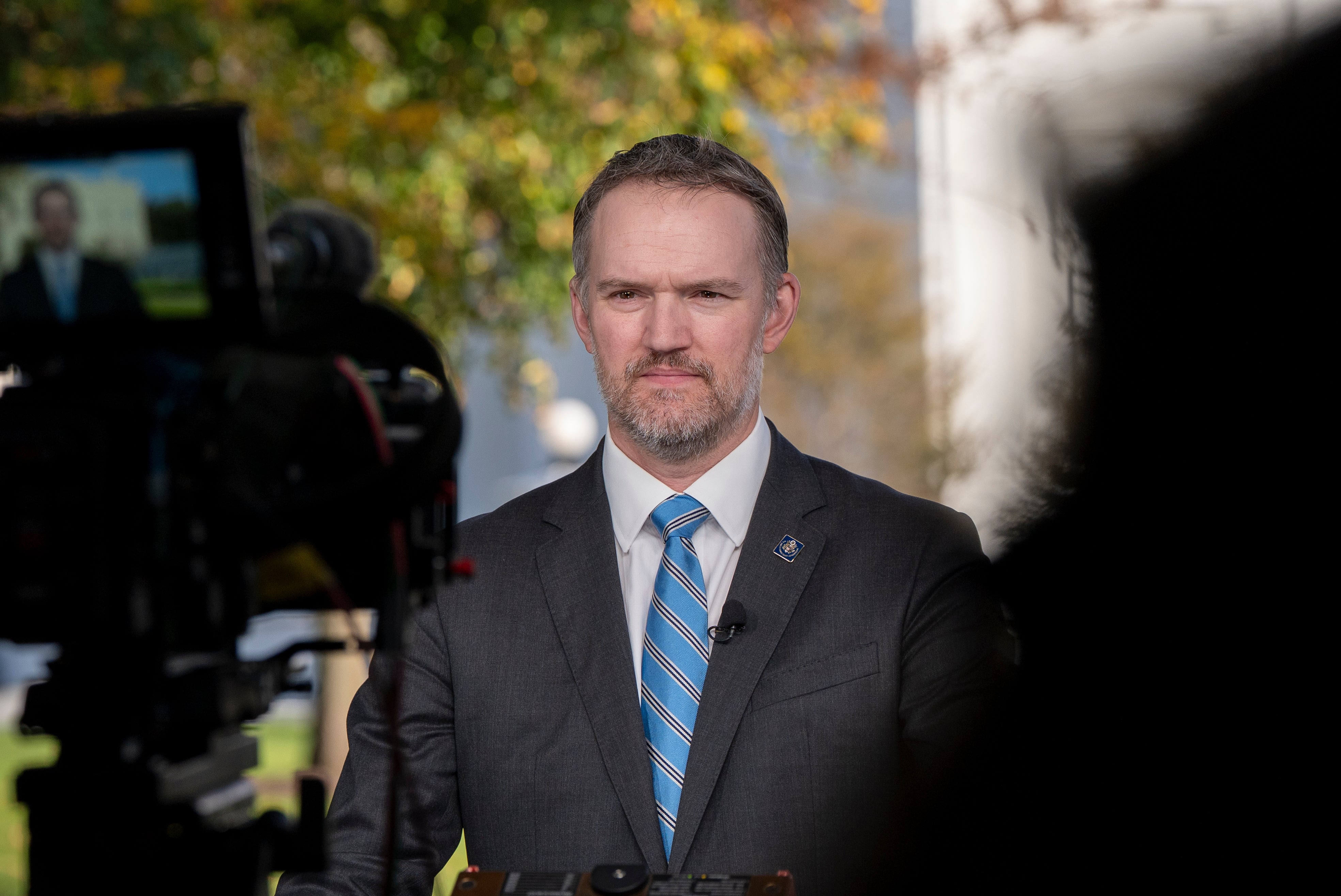 Man i mörkgrå kostym och blå slips intervjuas framför kamera utomhus med höstlöv i bakgrunden.