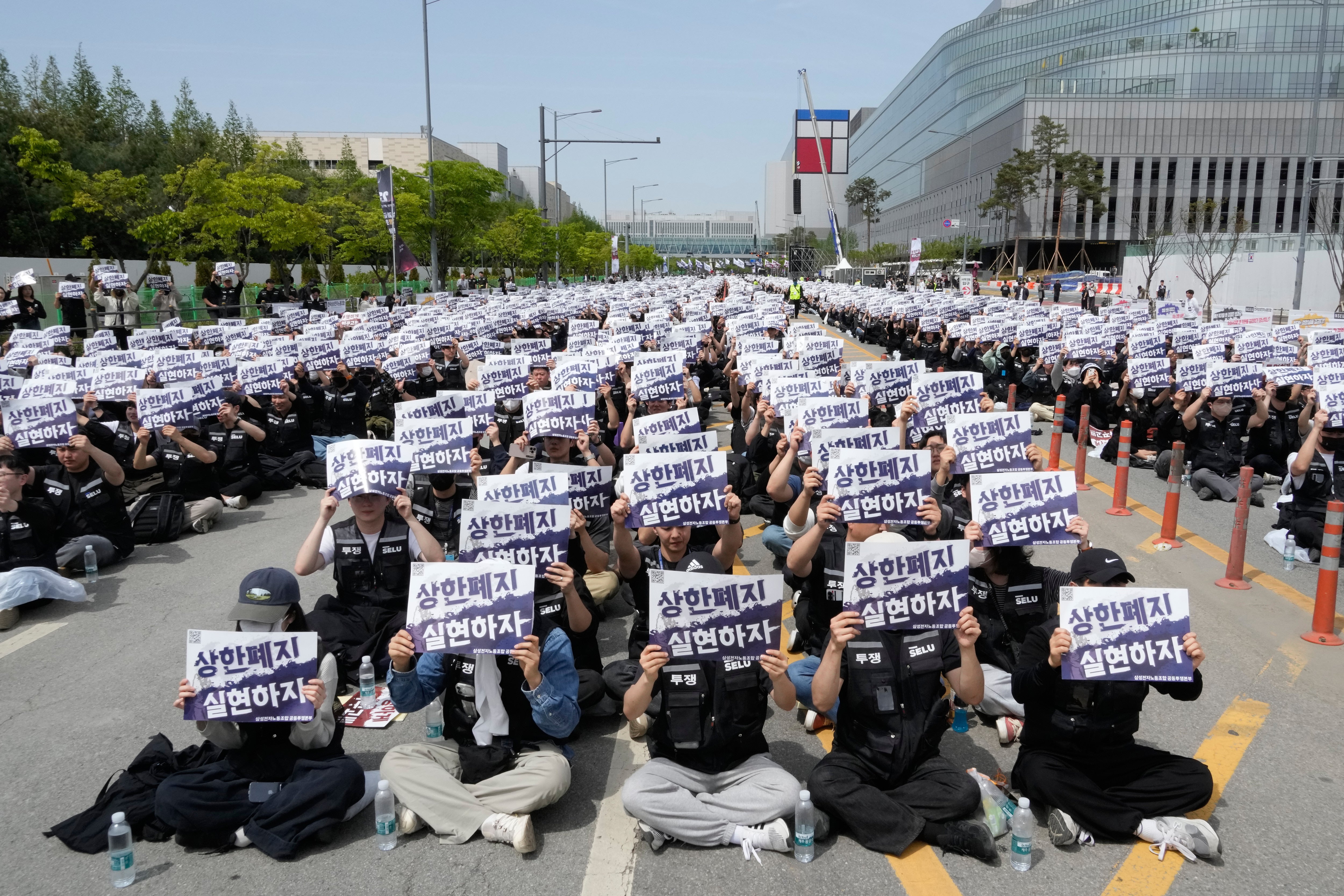 Stor demonstration med hundratals människor som sitter på en väg och håller upp vita protestskyltningar med koreansk text.