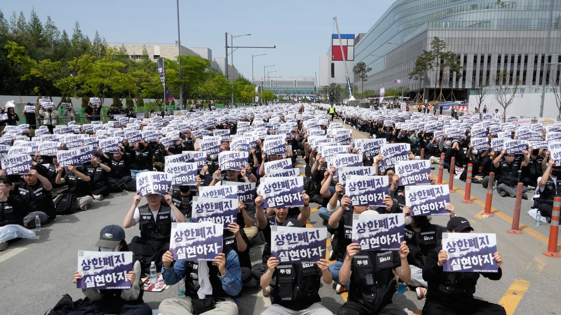 Stor demonstration med hundratals människor som sitter på en väg och håller upp vita protestskyltningar med koreansk text.