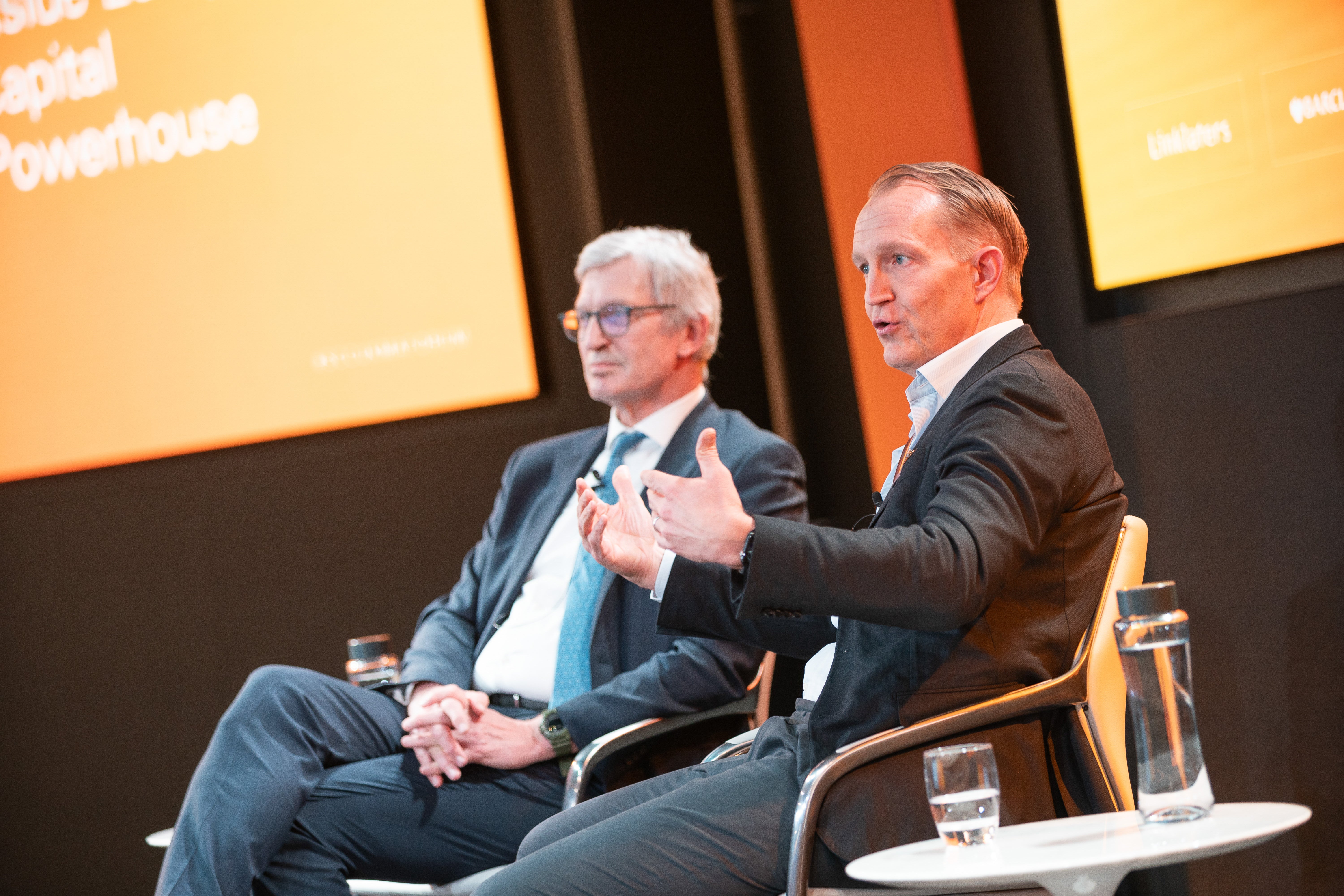 Två män i affärskläder diskuterar på en scen under en konferens.