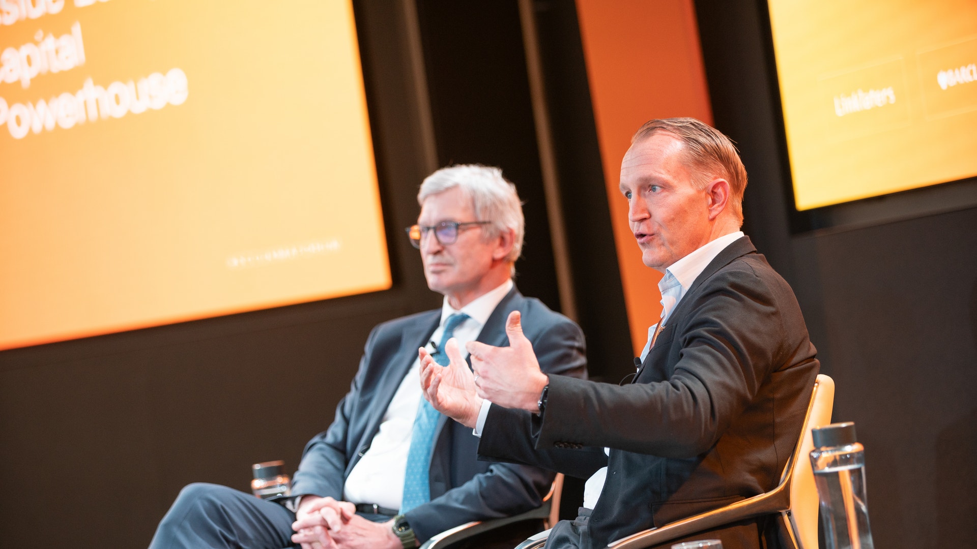 Två män i affärskläder diskuterar på en scen under en konferens.