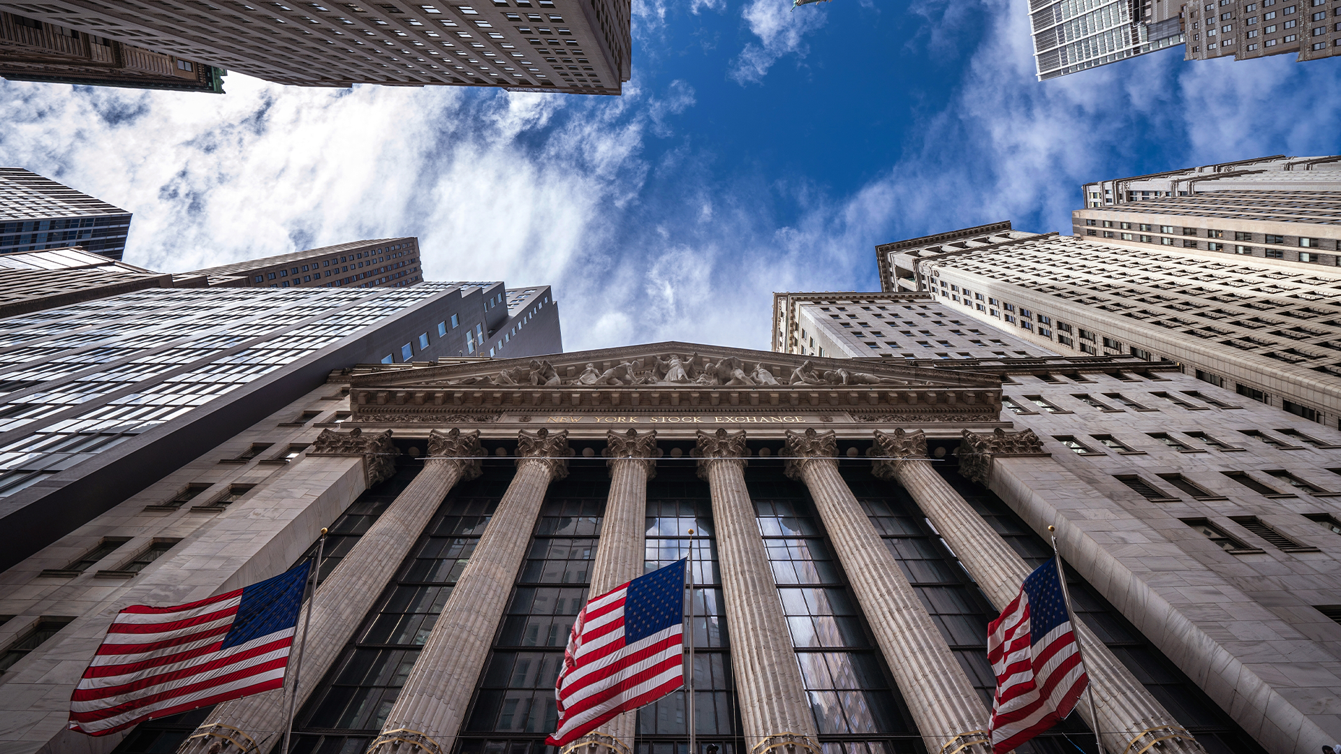 New York Stock Exchange-byggnaden fotograferad från nedan med höga kontorsbyggnader omkring och amerikanska flaggor framför fasaden.