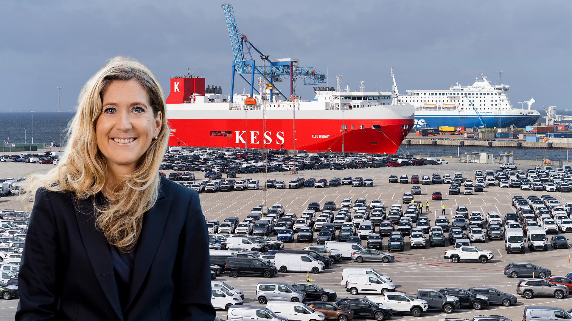 Leende kvinna med långt blont hår och mörk kavaj framför en hamn med många parkerade bilar och ett rött fartyg.