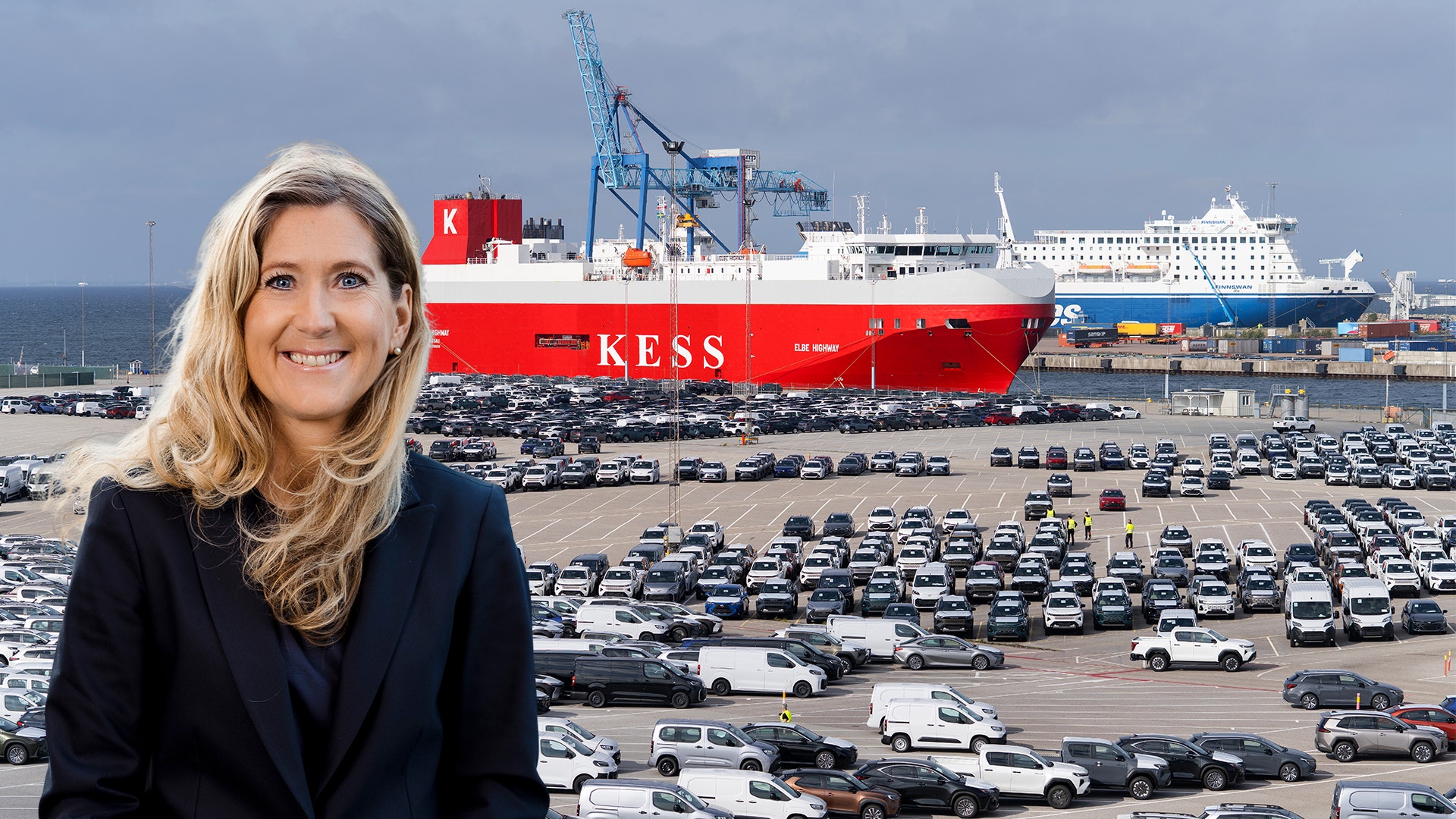 Leende kvinna med långt blont hår och mörk kavaj framför en hamn med många parkerade bilar och ett rött fartyg.