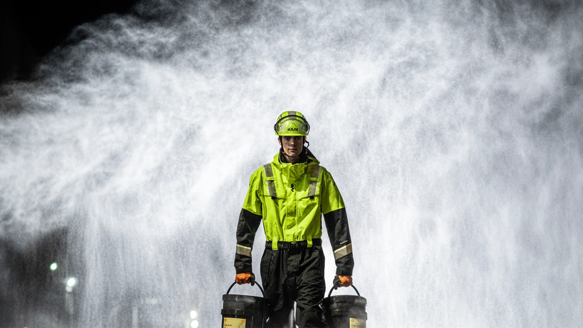 En person i varselkläder och hjälm står framför en stor vattenspray i mörkret.