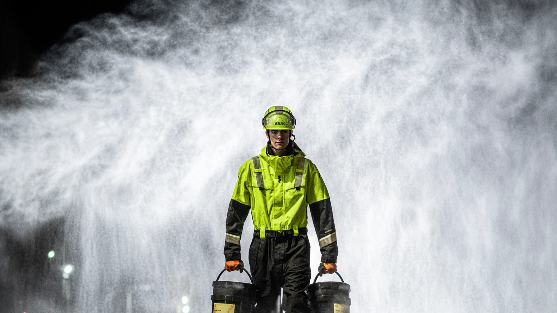 En person i varselkläder och hjälm står framför en stor vattenspray i mörkret.