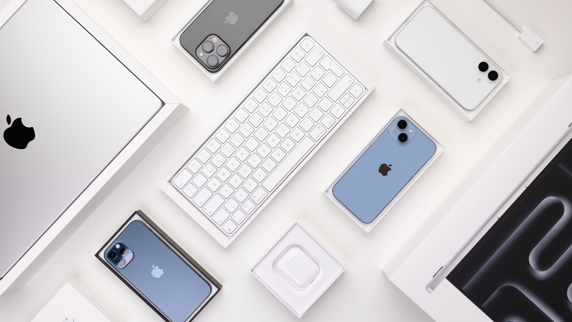 Samling av Apple-produkter och tillbehör arrangerade på vit bakgrund, inklusive MacBook, iPhone-telefoner, trådlöst tangentbord och AirPods-laddare.
