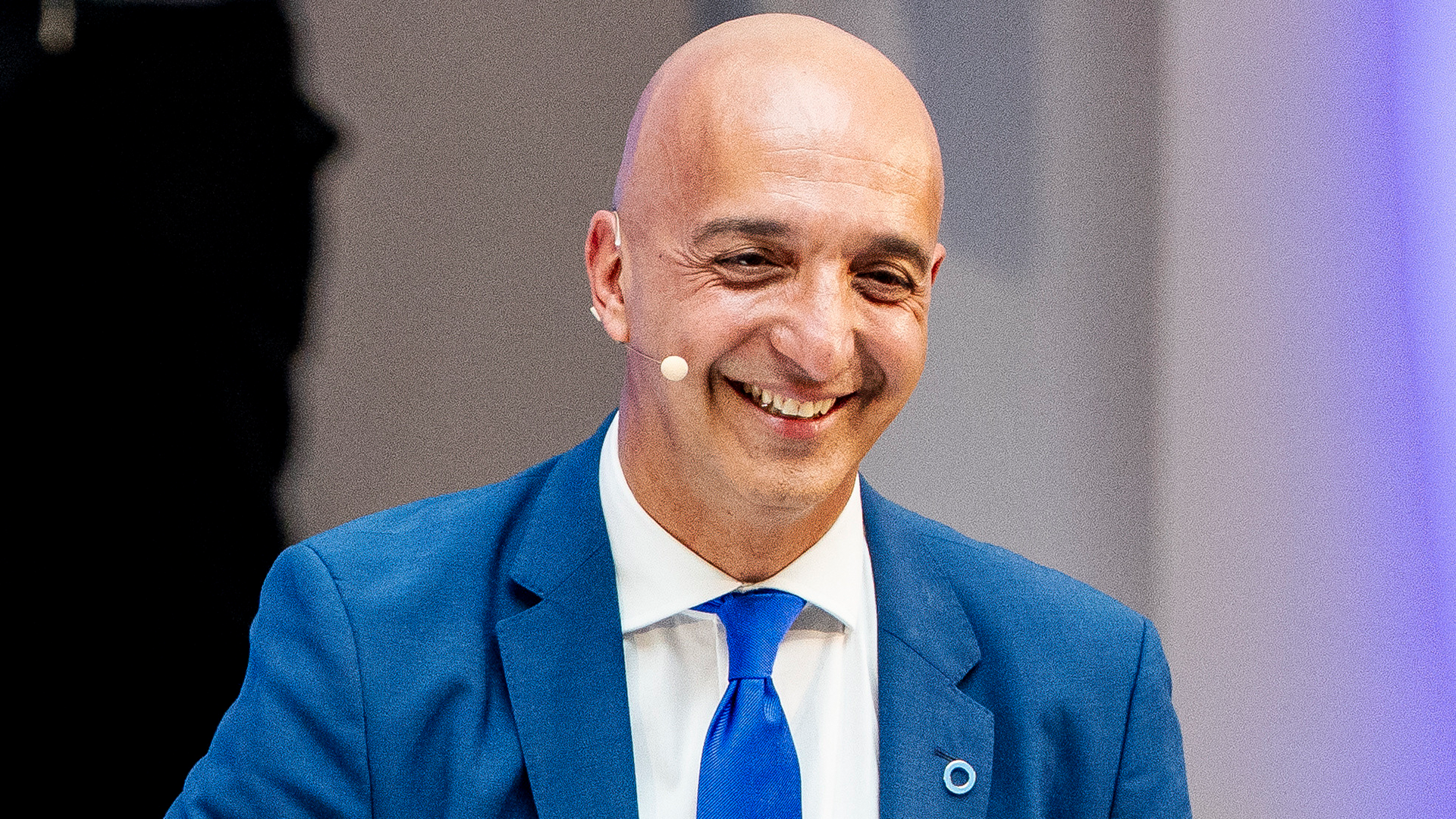 En leende man i kostym med slips talar med mikrofon på en scen.