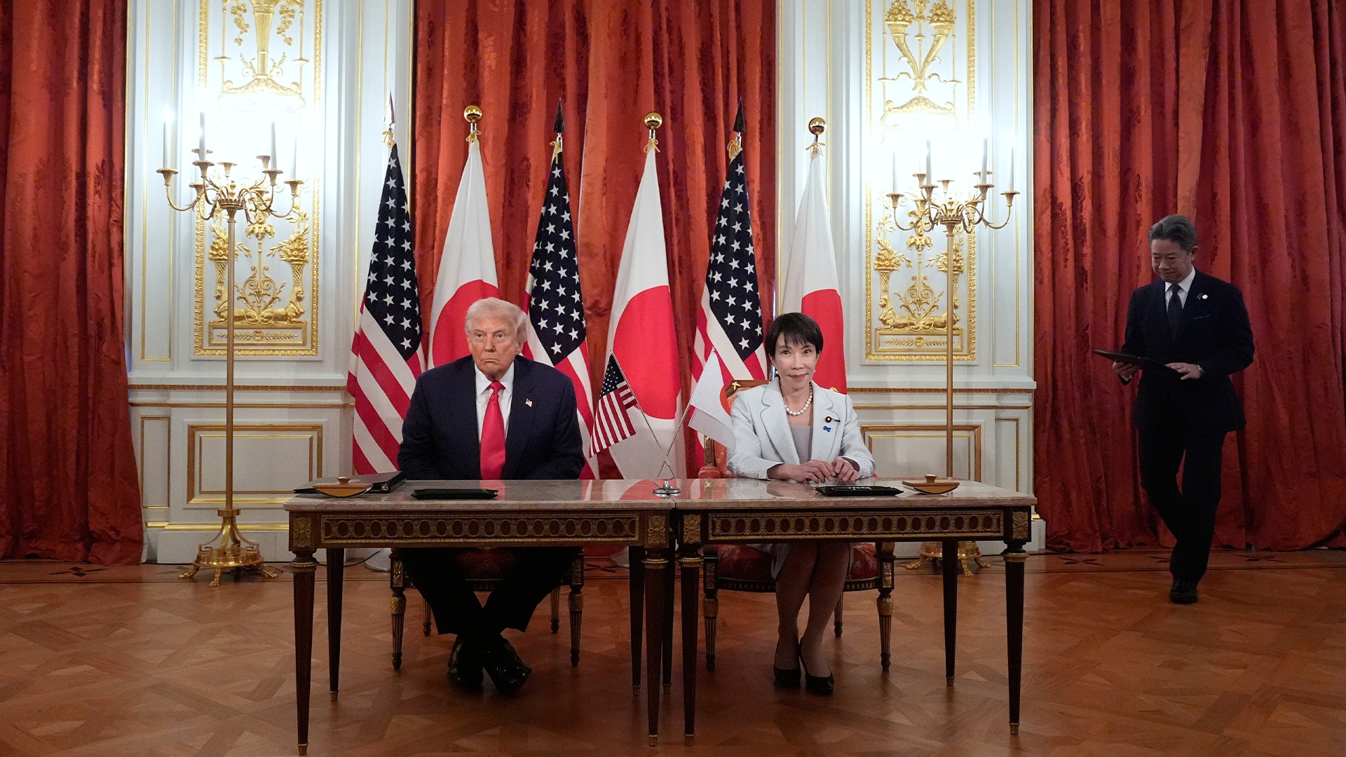 Två personer sitter vid ett bord under en officiell ceremoni med amerikanska, kanadensiska och japanska flaggor i bakgrunden. Rummet är elegant inrett med röda gardiner och gyllene ljusstakar.