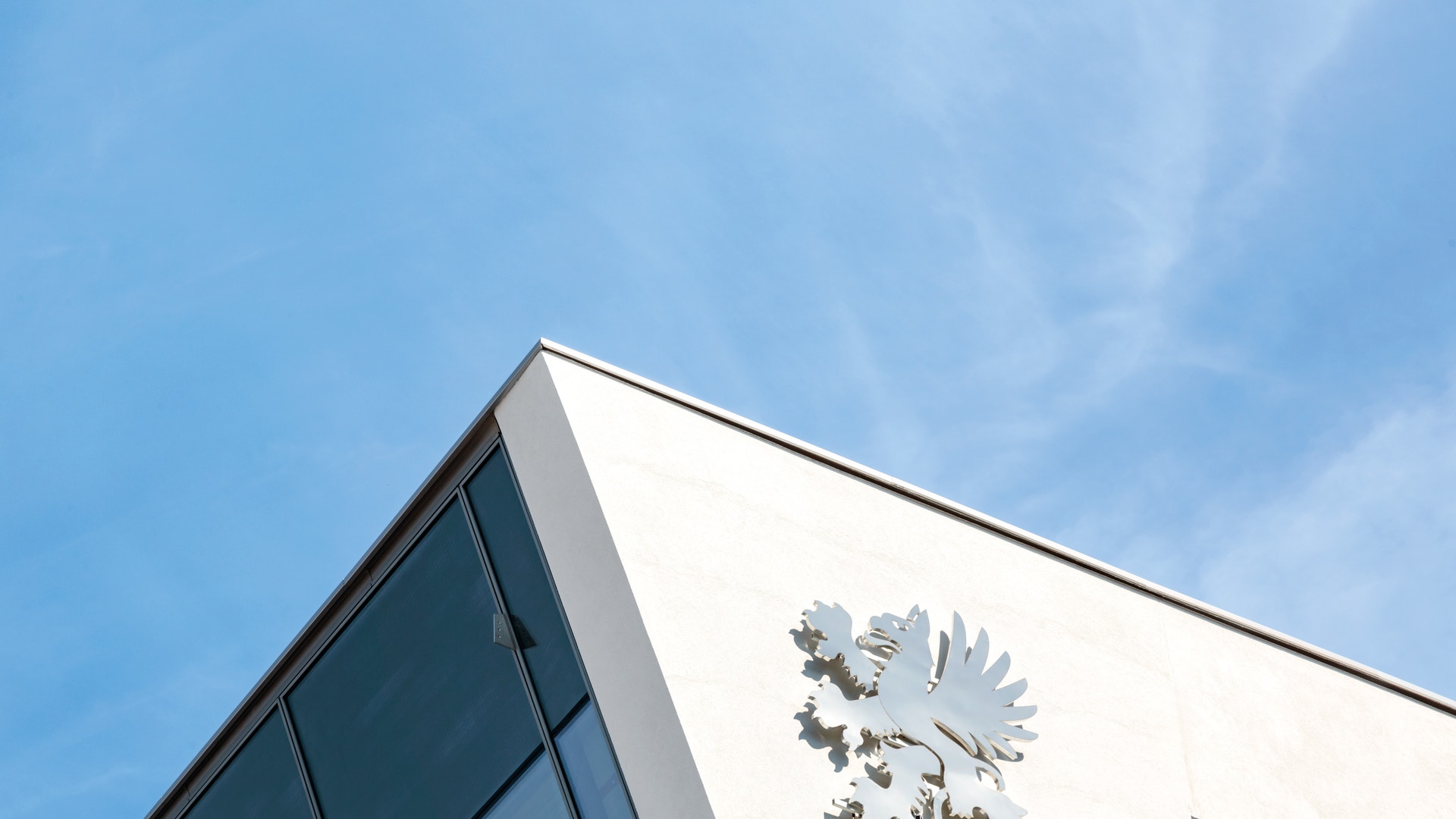 Hörnet på en ljus byggnad med mörka fönster och en vit grip-skulptur på fasaden, sedd underifrån mot en blå himmel.