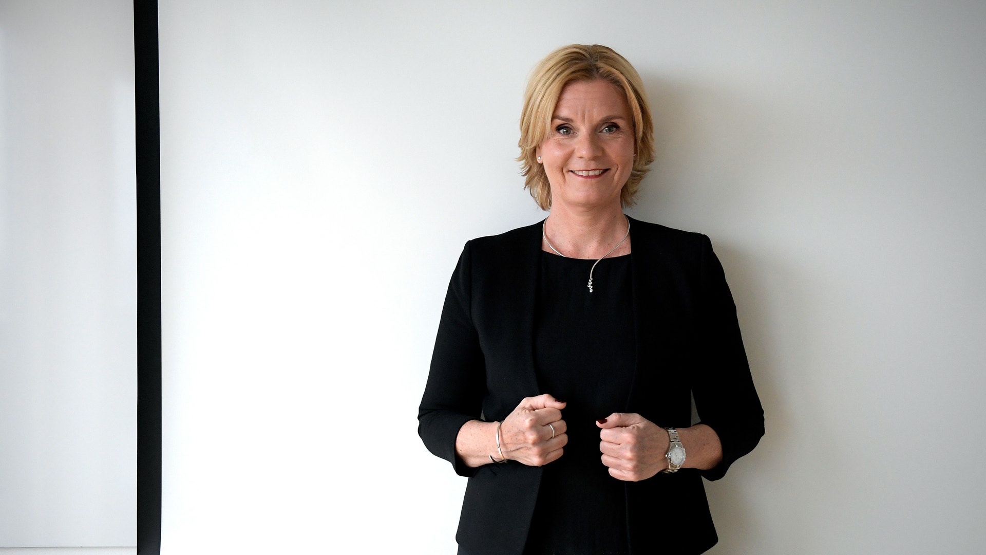 Leende kvinna med kort ljust hår och svart kavaj står med knutna händer framför en vit projektorduk.