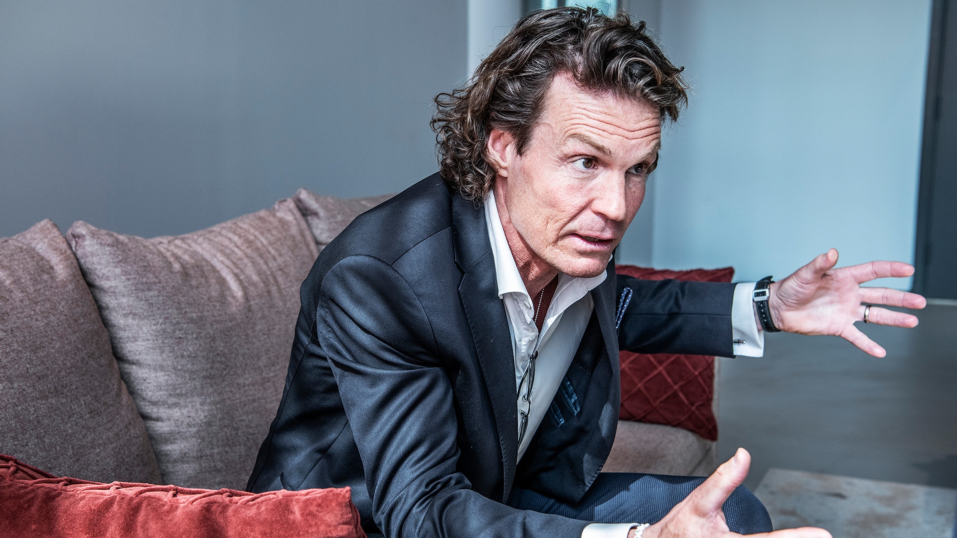 Man i svart kostym sitter på soffa och gestikulerar medan han talar.