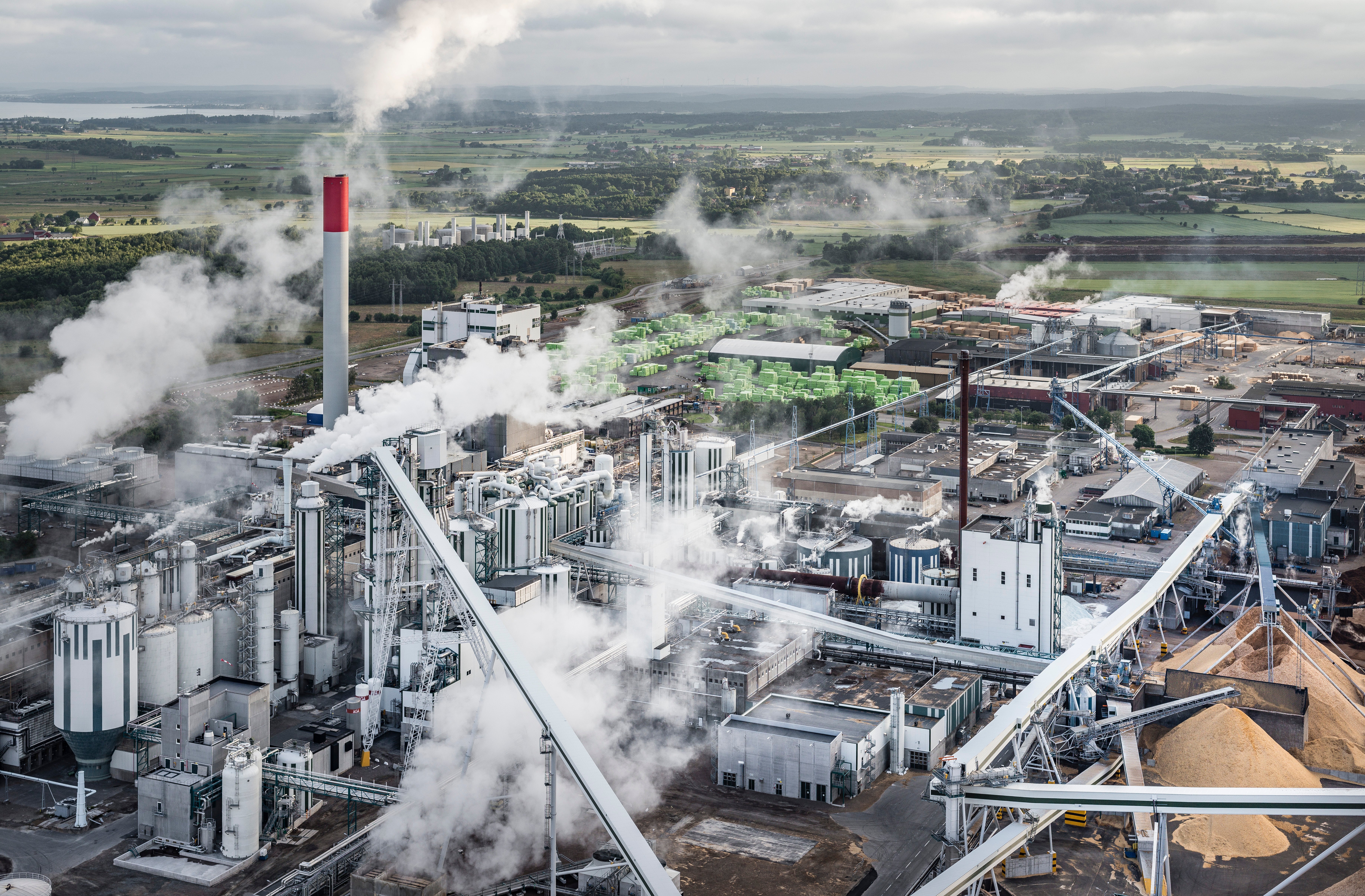 Flygfoto över en stor industrianläggning med fabriksbyggnader, skorstenar och rökutsläpp. Anläggningen omges av åkermark och gröna fält.