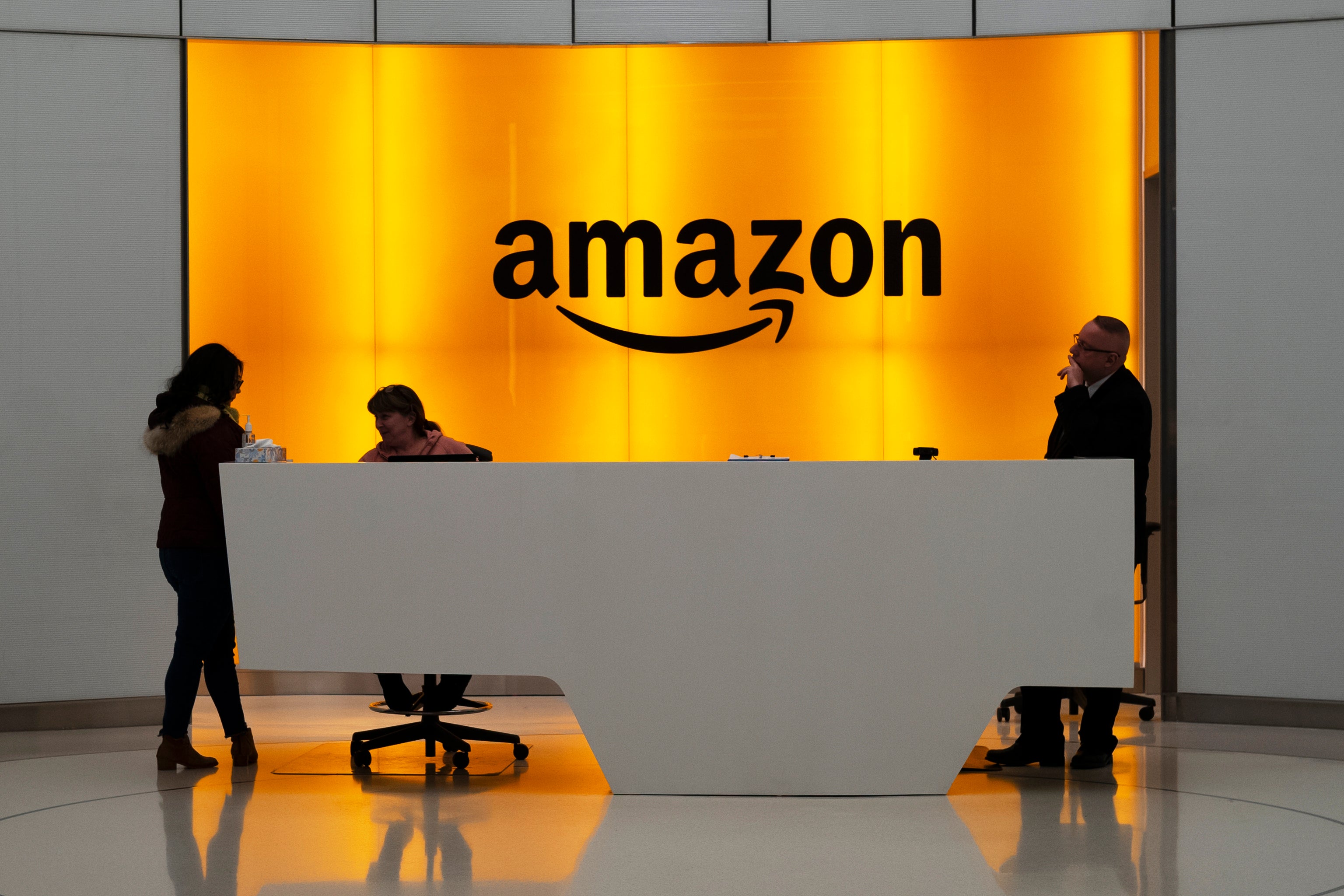 Amazons logotyp på en upplyst orange vägg bakom en reception, där några personer står och sitter.