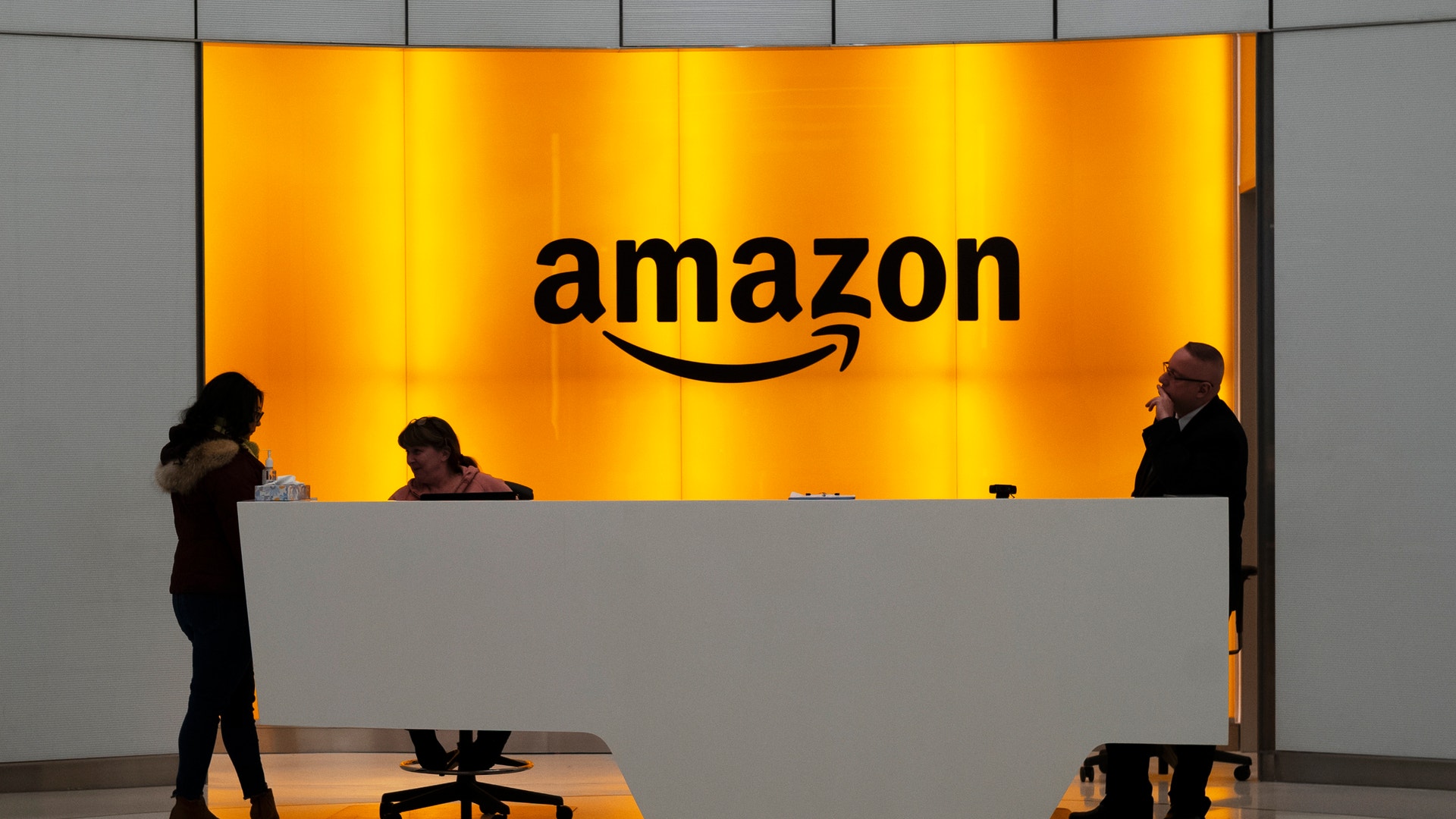 Amazons logotyp på en upplyst orange vägg bakom en reception, där några personer står och sitter.