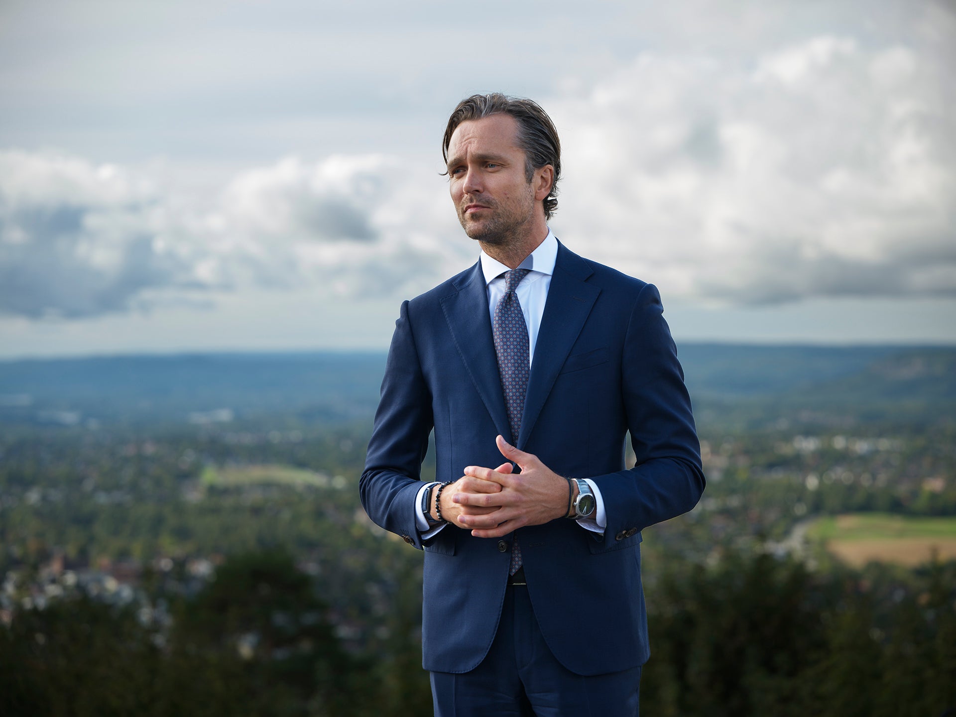 En man i kostym står utomhus med utsikt över en stad och kullar i bakgrunden.