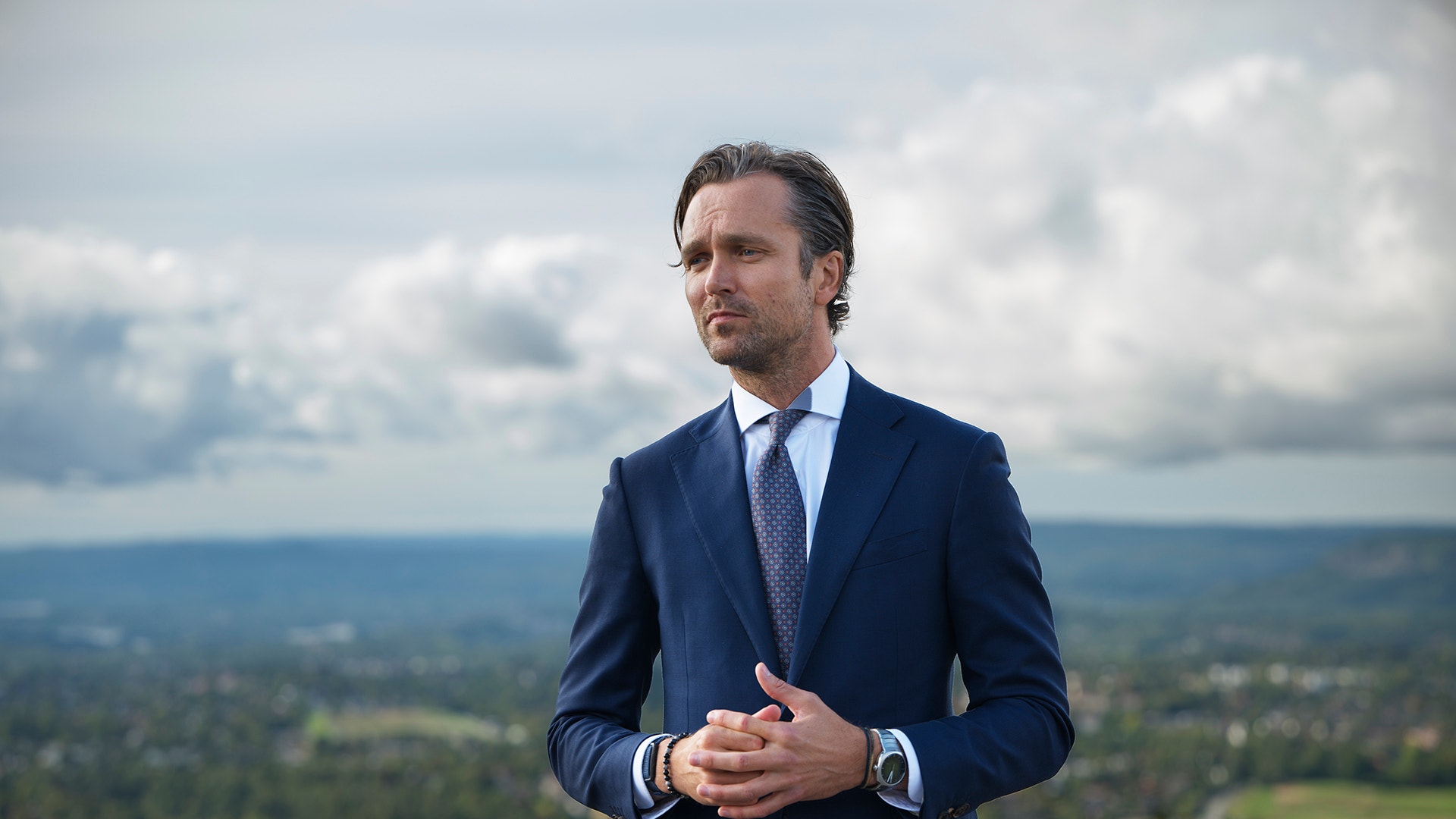 En man i kostym står utomhus med utsikt över en stad och kullar i bakgrunden.