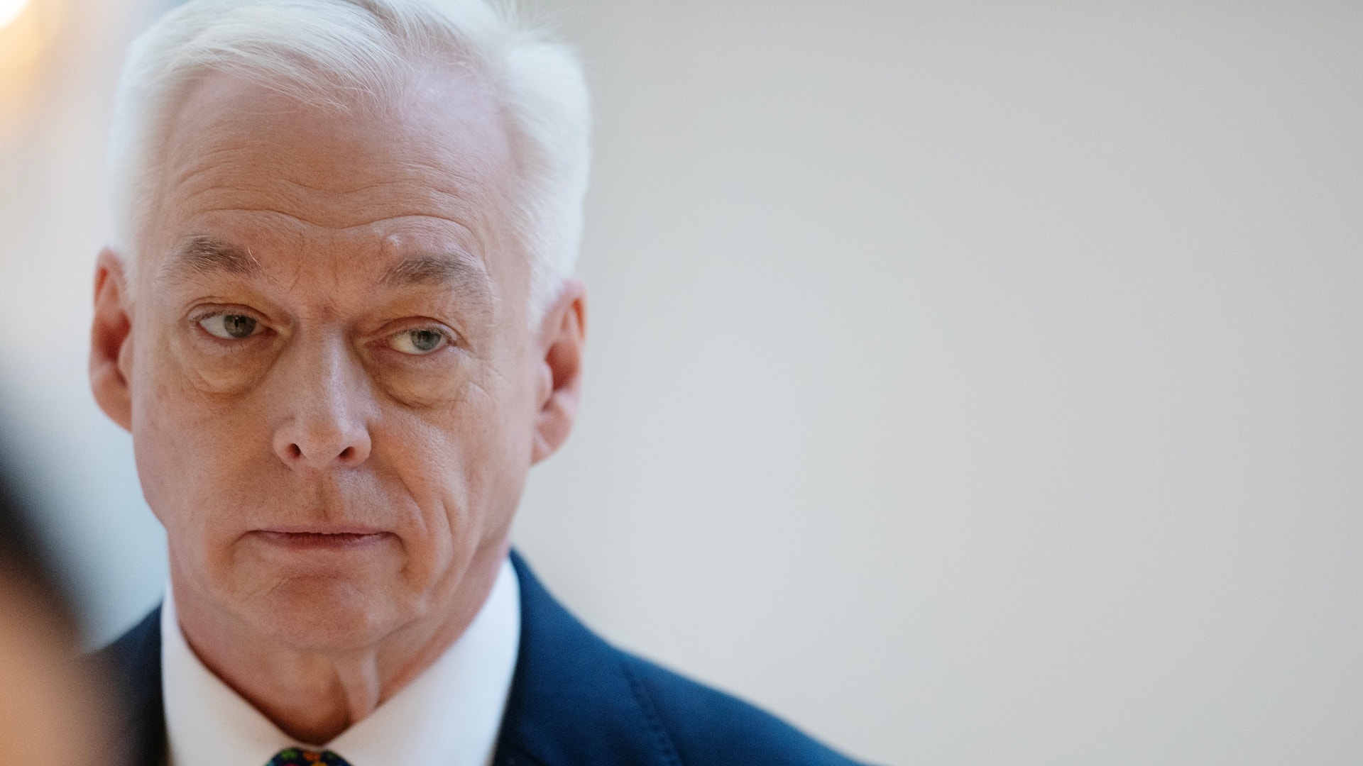 Porträttfoto av en äldre man med vitt hår, iklädd mörkblå kostym och färgglad slips.