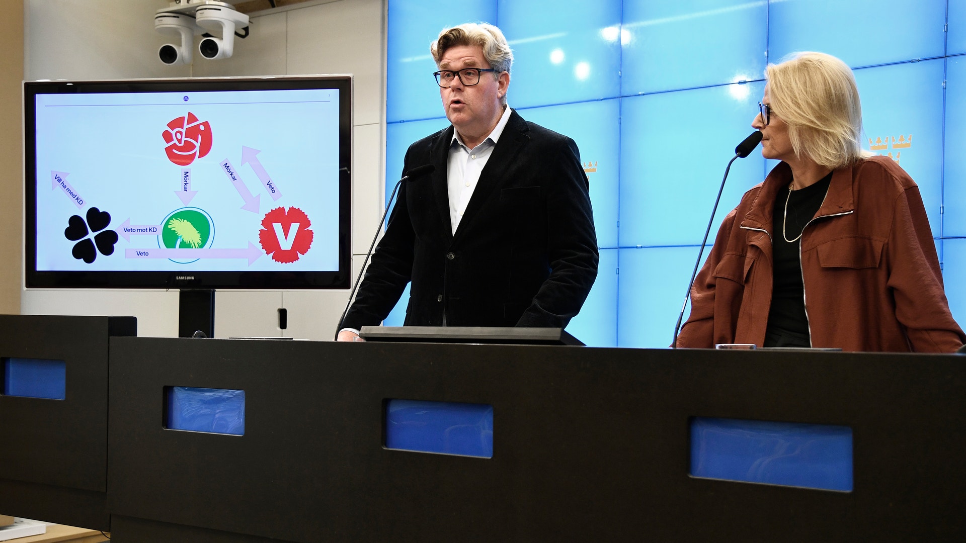 En man och en kvinna presenterar vid ett podium framför en skärm som visar ett diagram med logotyper och symboler.