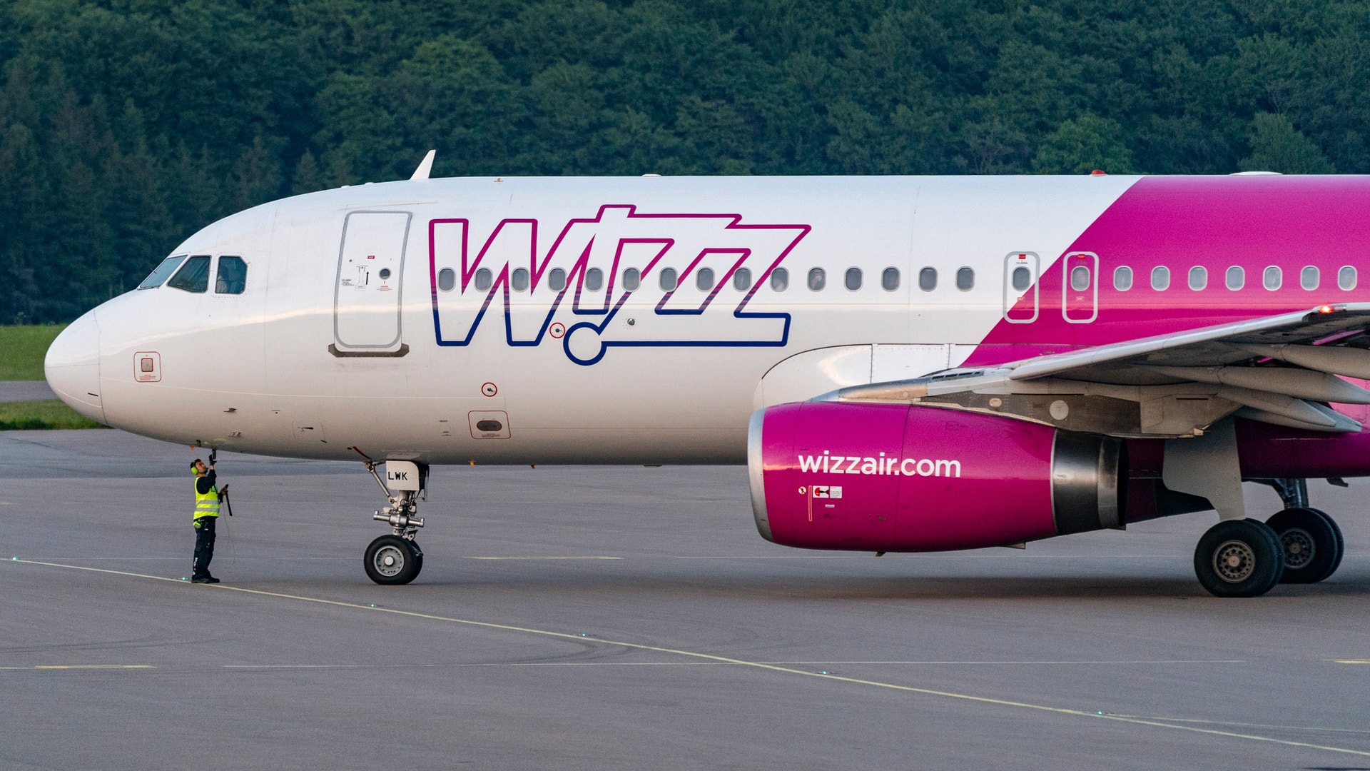 Wizz Air-flygplan parkerat på landningsbanans betongyta med en markarbetare i gul väst i närheten.