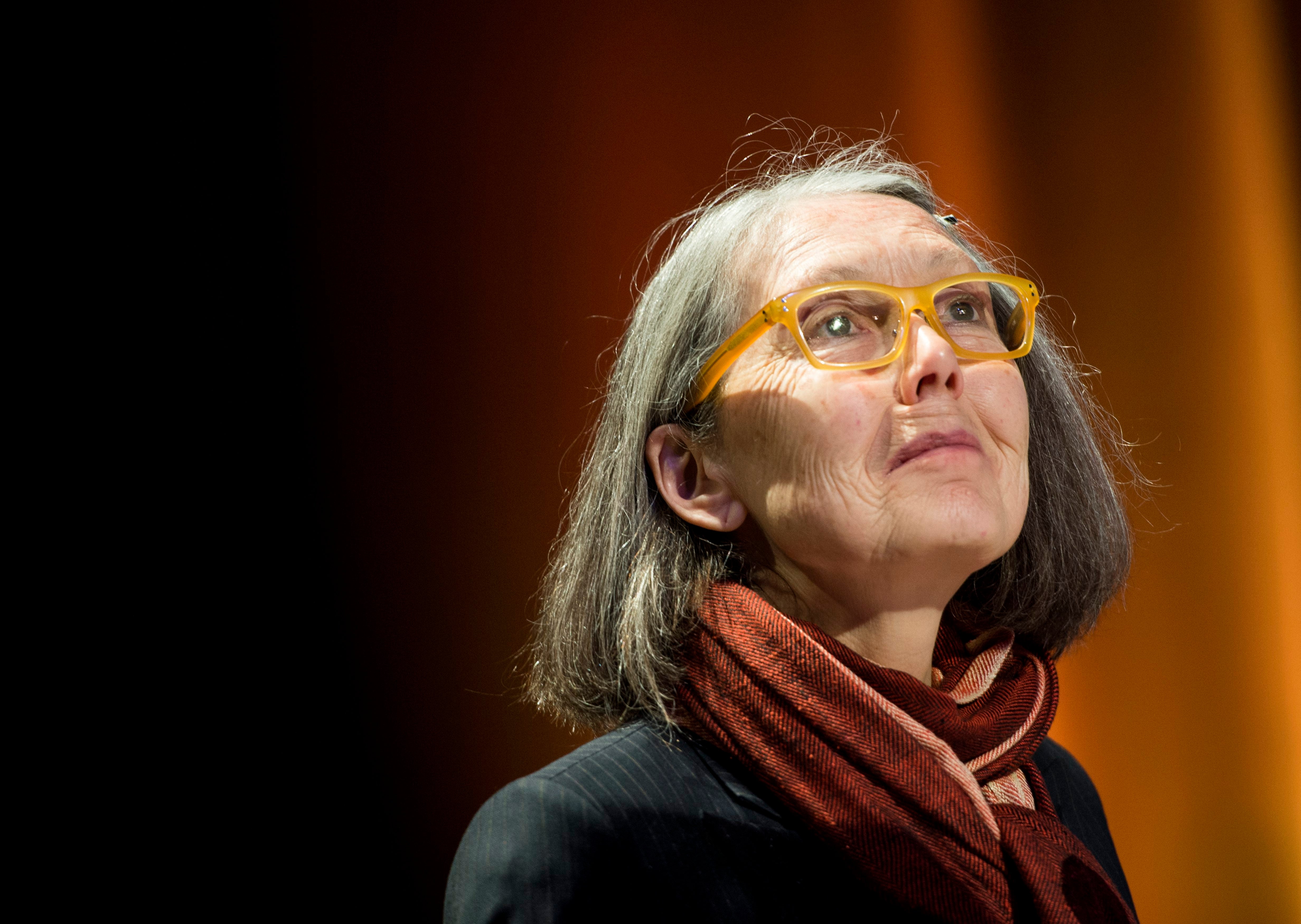 Poeten Anne Carson med grått hår, gula glasögon och rödbrun halsduk blickar uppåt mot en mörk scen.