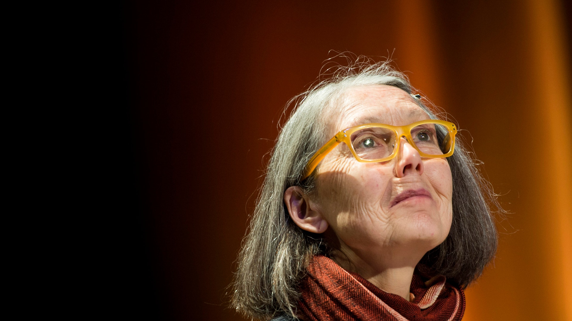 Poeten Anne Carson med grått hår, gula glasögon och rödbrun halsduk blickar uppåt mot en mörk scen.
