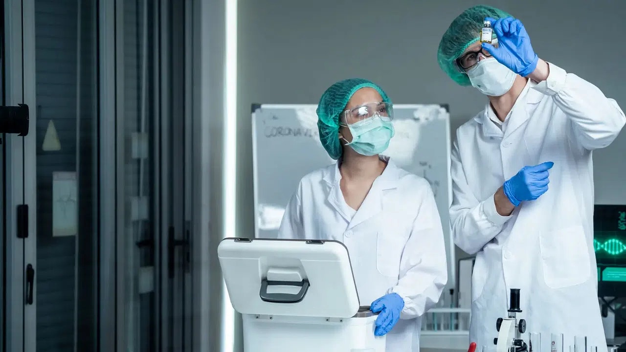 Två forskare i skyddskläder och munskydd arbetar i ett laboratorium, en av dem håller upp en liten flaska.
