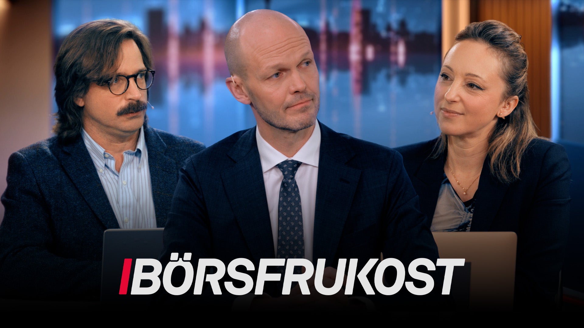 Tre personer i affärskläder sitter tillsammans i en TV-studio med stadsvy i bakgrunden.