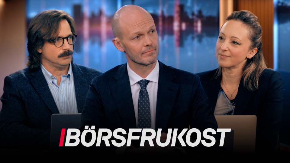 Tre personer i affärskläder sitter tillsammans i en TV-studio med stadsvy i bakgrunden.