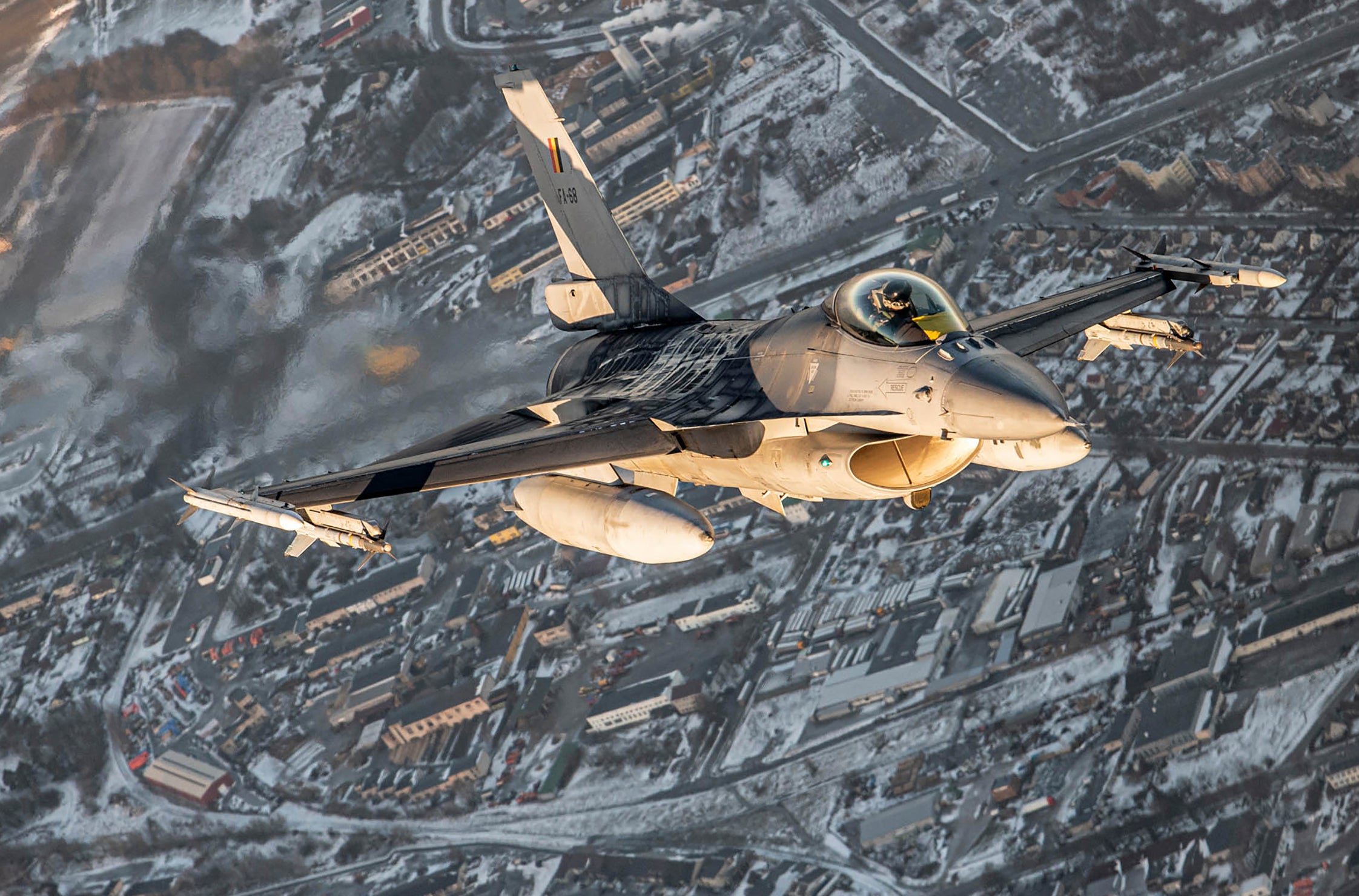 Luftfoto av ett militärt stridsflygplan i flight över snöig bergsterräng med vägar och bebyggelse synlig under planet.