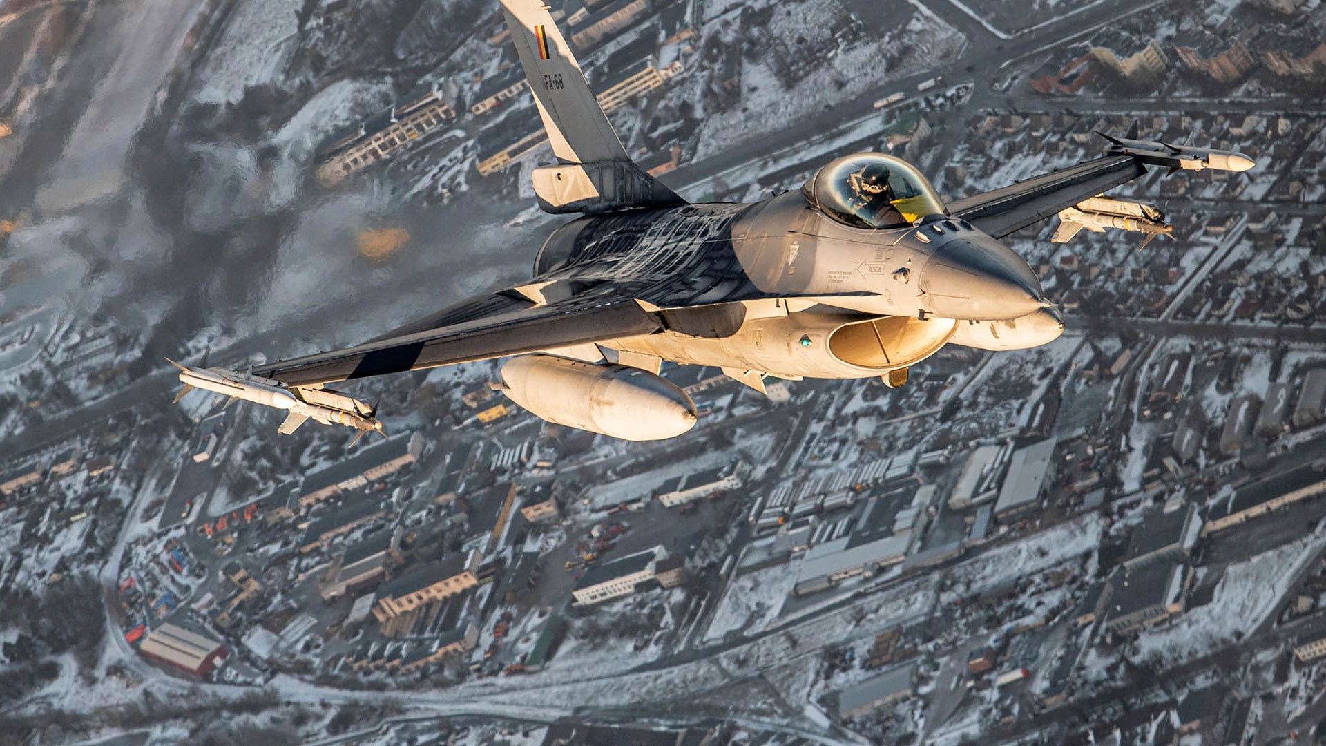 Luftfoto av ett militärt stridsflygplan i flight över snöig bergsterräng med vägar och bebyggelse synlig under planet.