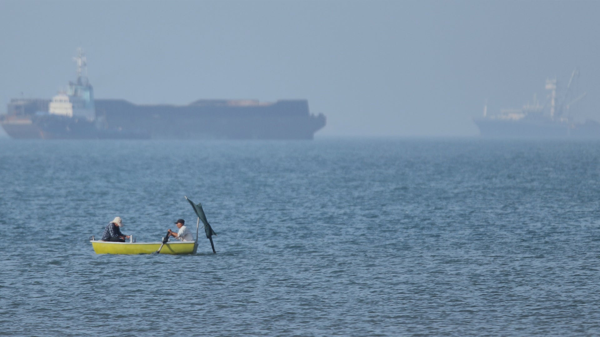 Två personer paddlar i en gul kajak på havet. I bakgrunden syns stora lastfartyg och industrianläggningar i dimman.