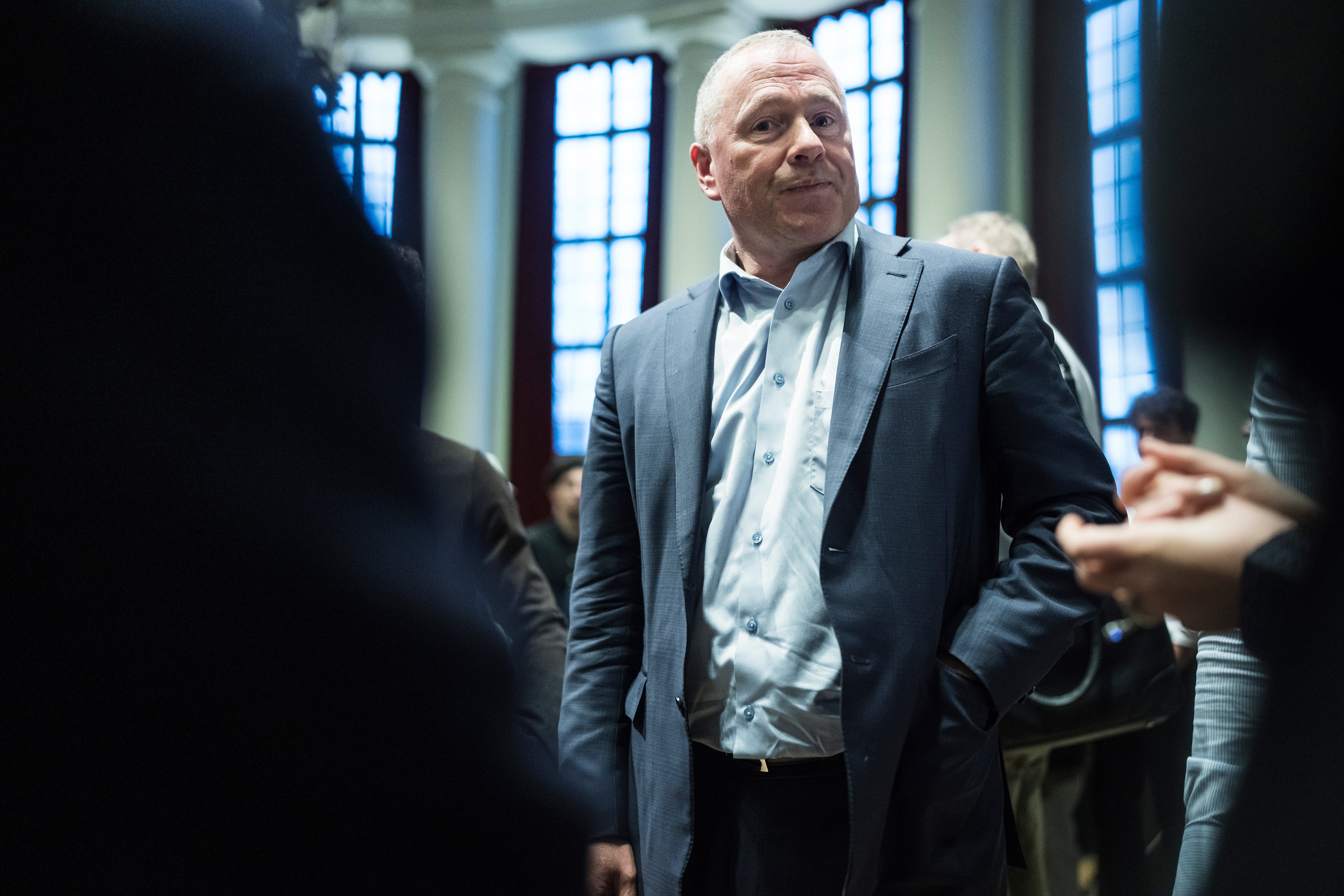 Man i mörk kostym och ljus skjorta står i ett rum med stora fönster, omgiven av andra personer.