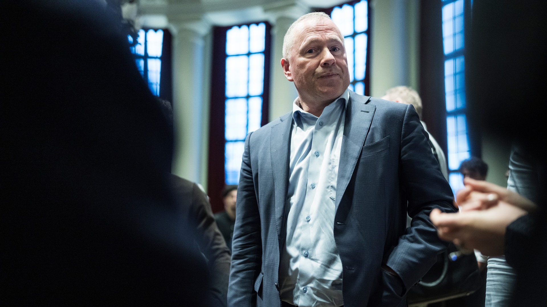 Man i mörk kostym och ljus skjorta står i ett rum med stora fönster, omgiven av andra personer.