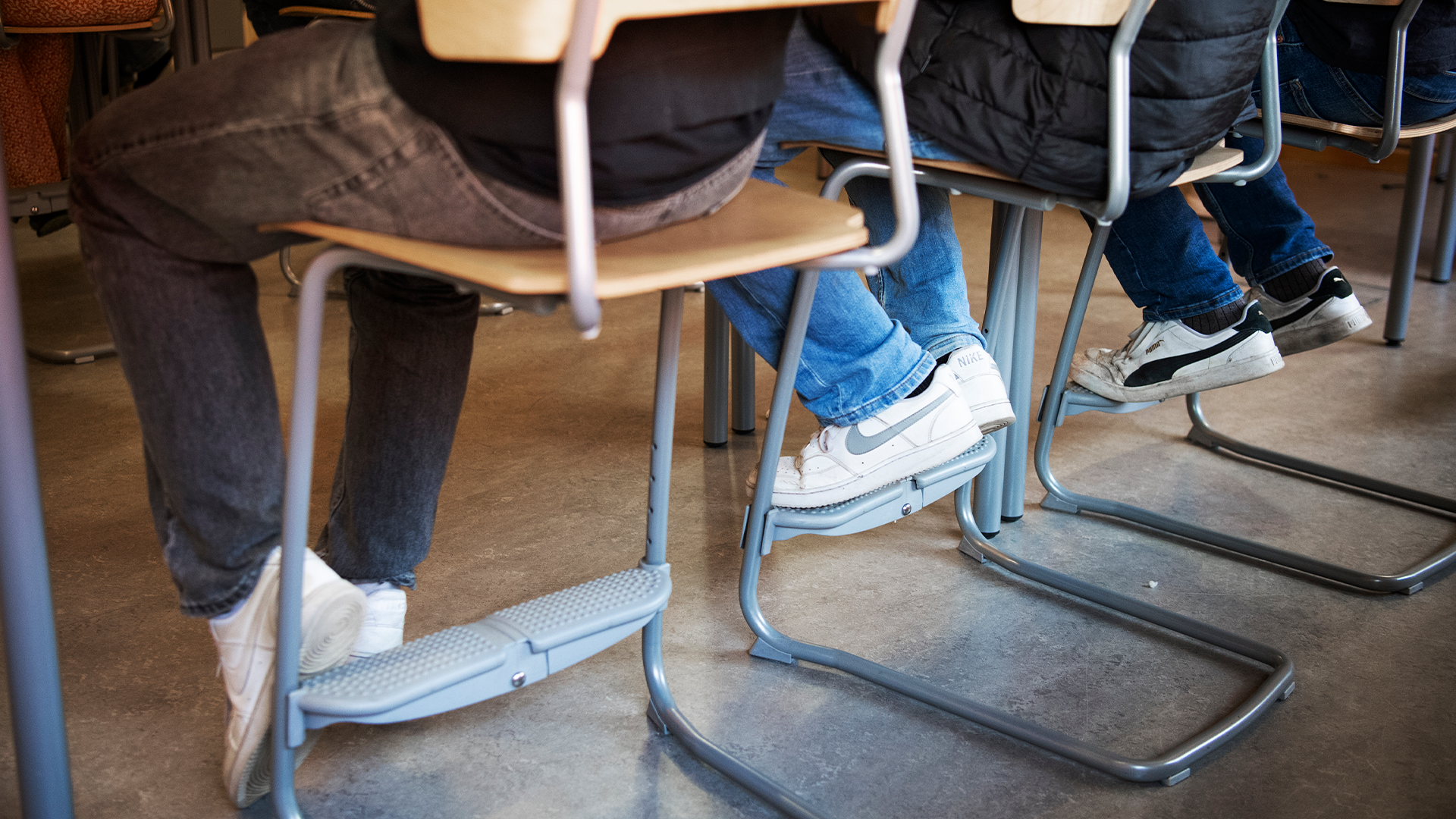 Elever sitter på stolar i ett klassrum, fotografi från lågt perspektiv som visar deras fötter och ben.