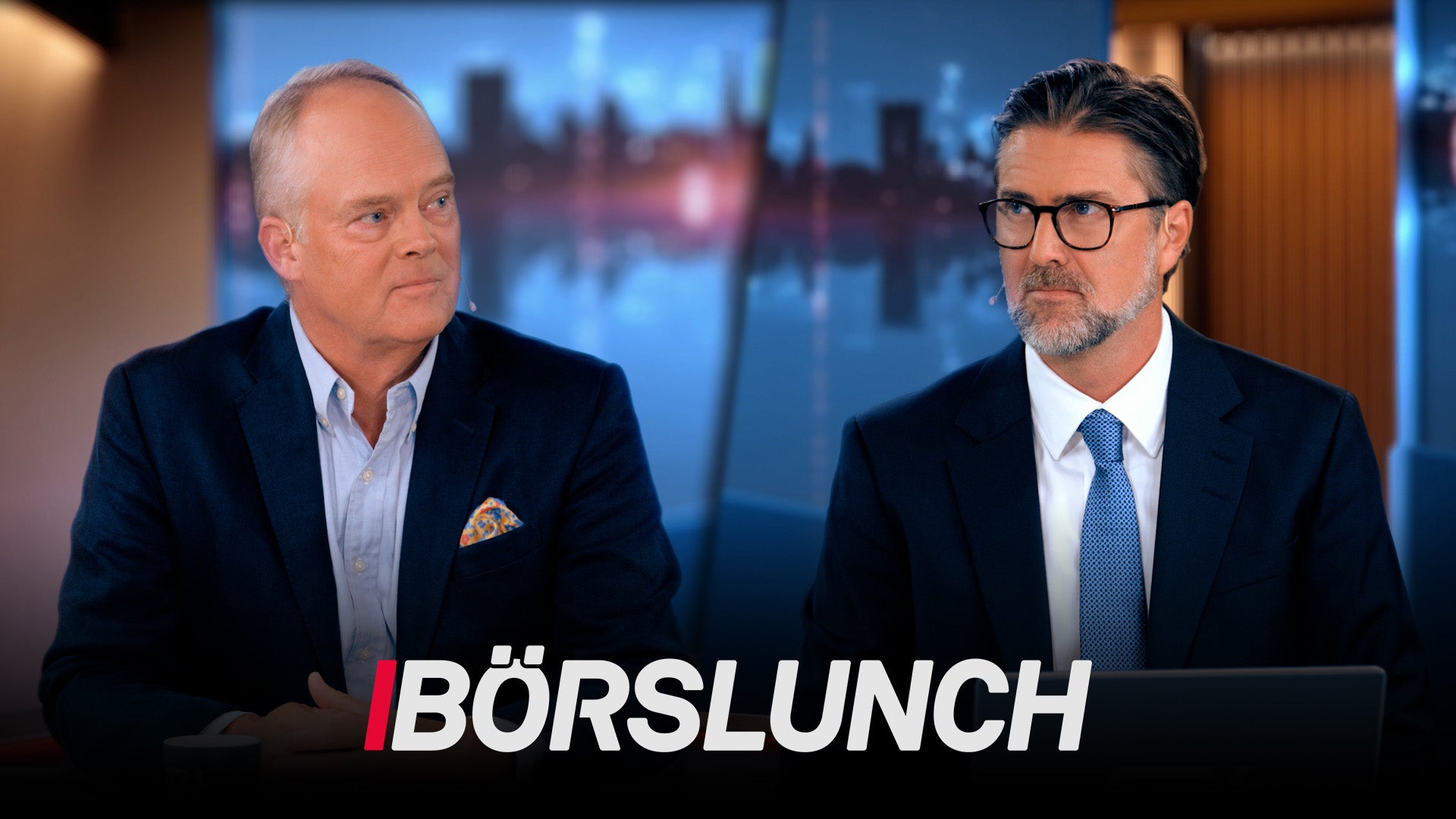 Två män i affärskläder sitter vid ett skrivbord under inspelning av TV-programmet Börslunch. Stadshorisont syns genom fönstret bakom dem.