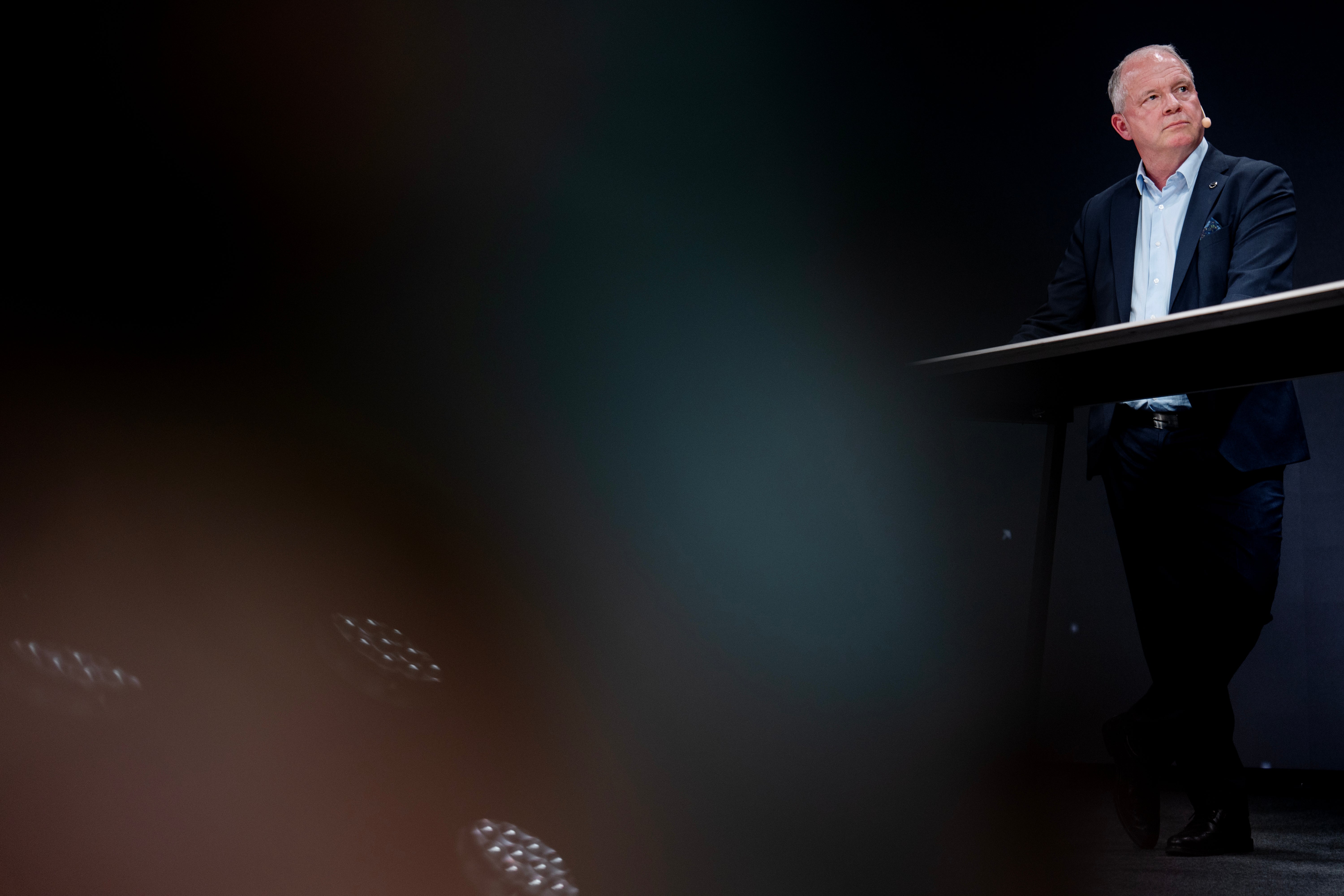 En man i mörk kostym talar vid ett podium på en scen framför en publik.