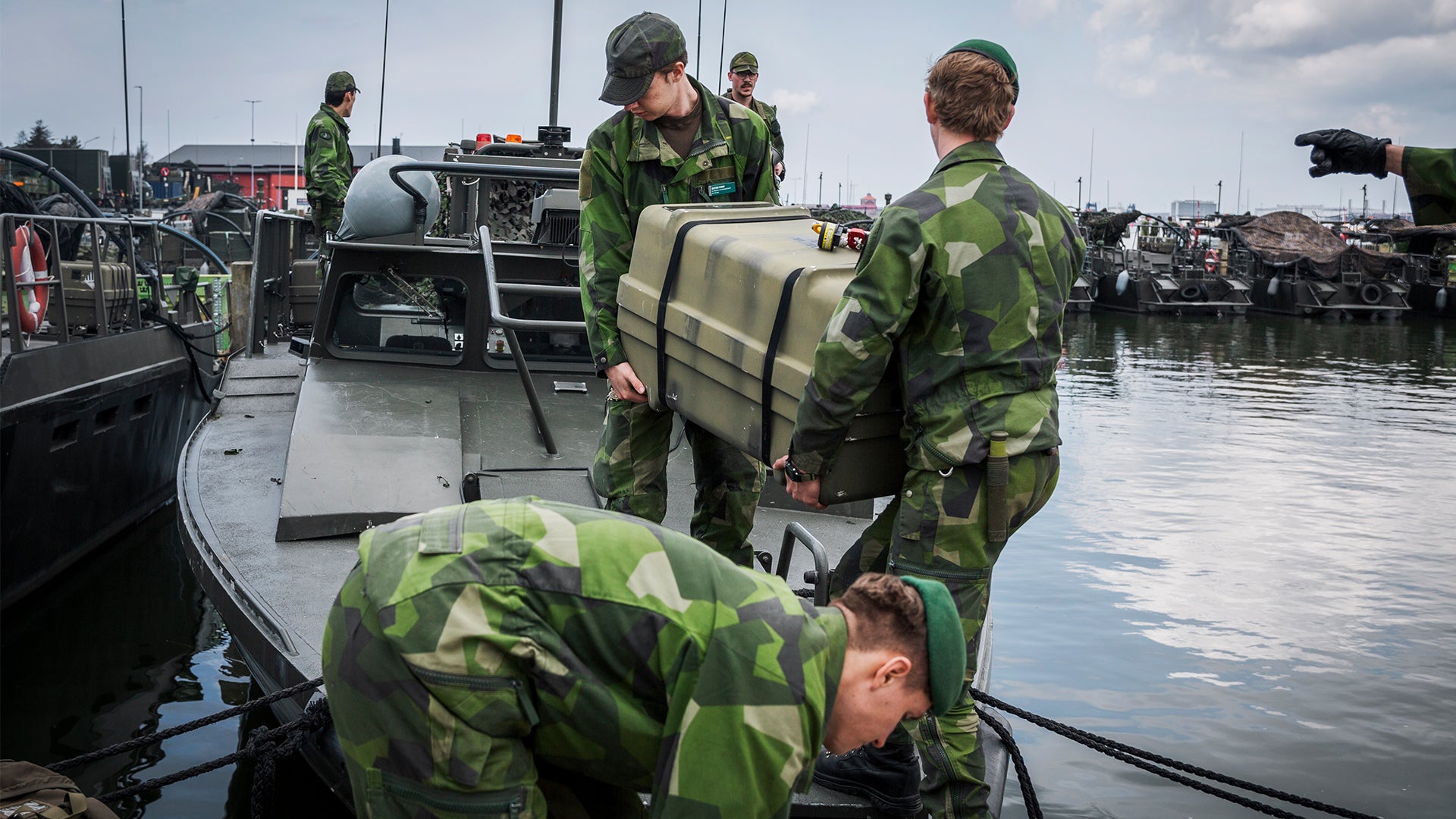 Svenska militärer i kamouflageuniformer arbetar vid militära båtar i en hamn. Soldaterna hanterar utrustning och förberedelser på båtarna.