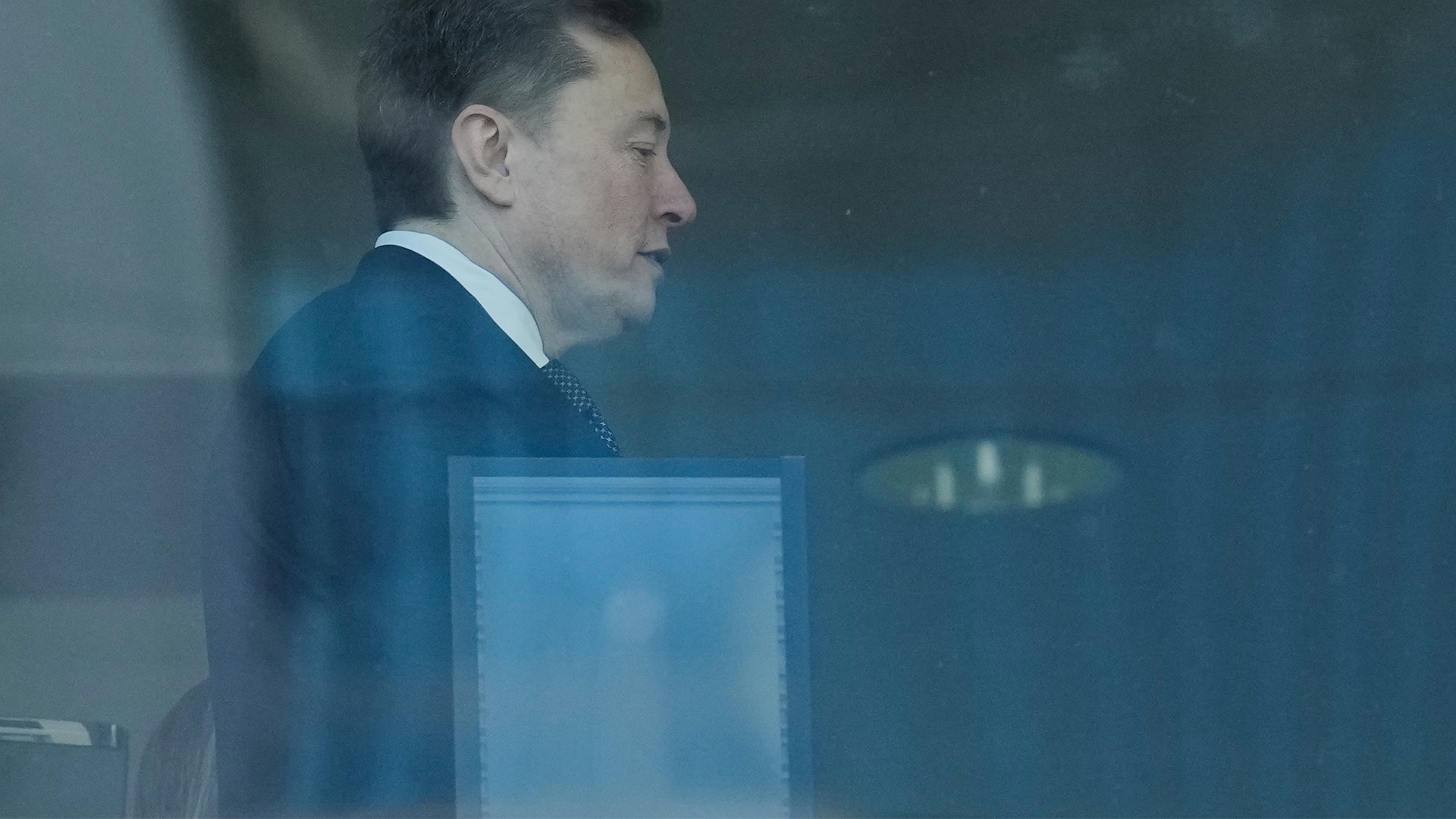 Man i blå kostym sitter bakom glas och tittar åt sidan.