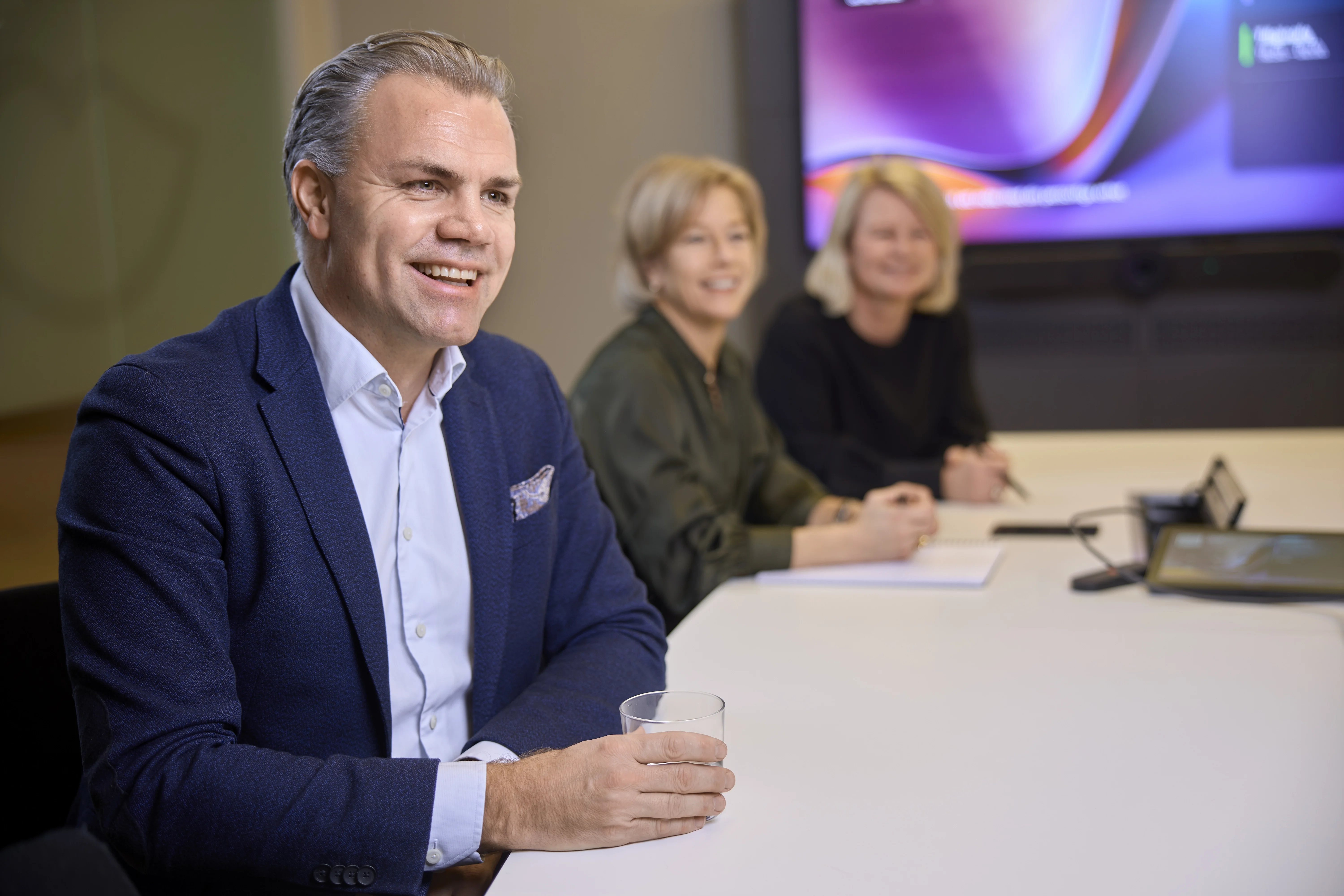 Man i mörk blazer vid ett bord under en professionell paneldiskussion med två kvinnor i bakgrunden.