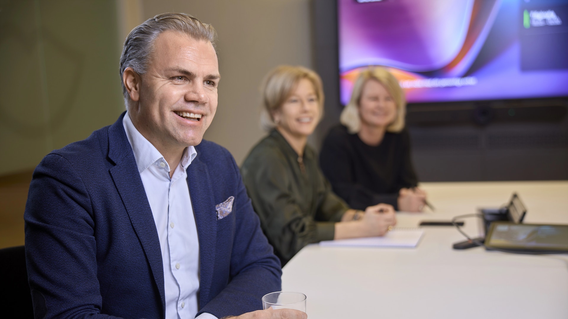Man i mörk blazer vid ett bord under en professionell paneldiskussion med två kvinnor i bakgrunden.
