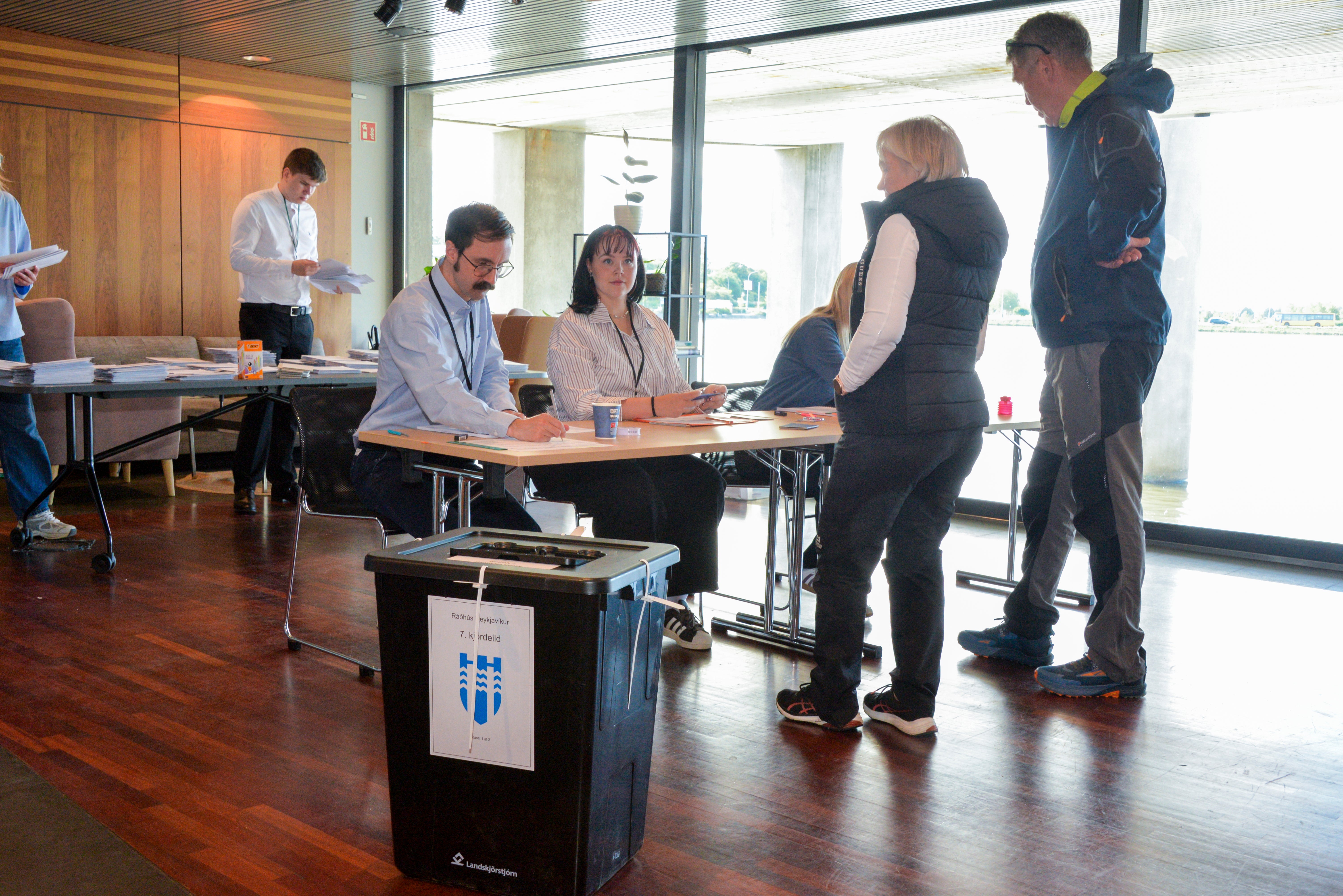 Flera personer samlade vid ett bord under ett möte eller arrangemang inomhus. En skräpkorg står i förgrunden och stora fönster syns i bakgrunden.