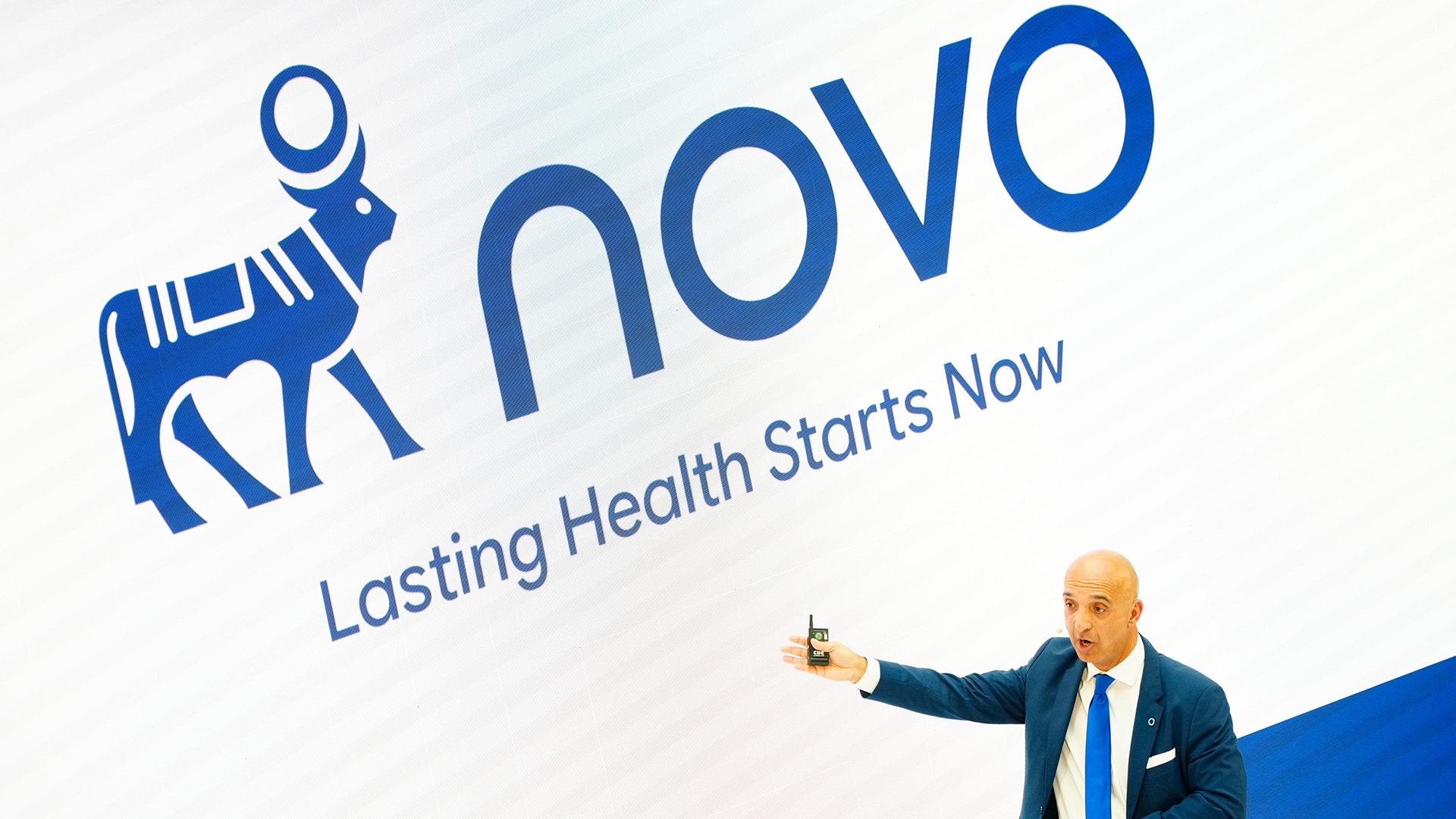 En man i kostym håller en presentation framför en skärm med Novos logotyp och texten "Lasting Health Starts Now".