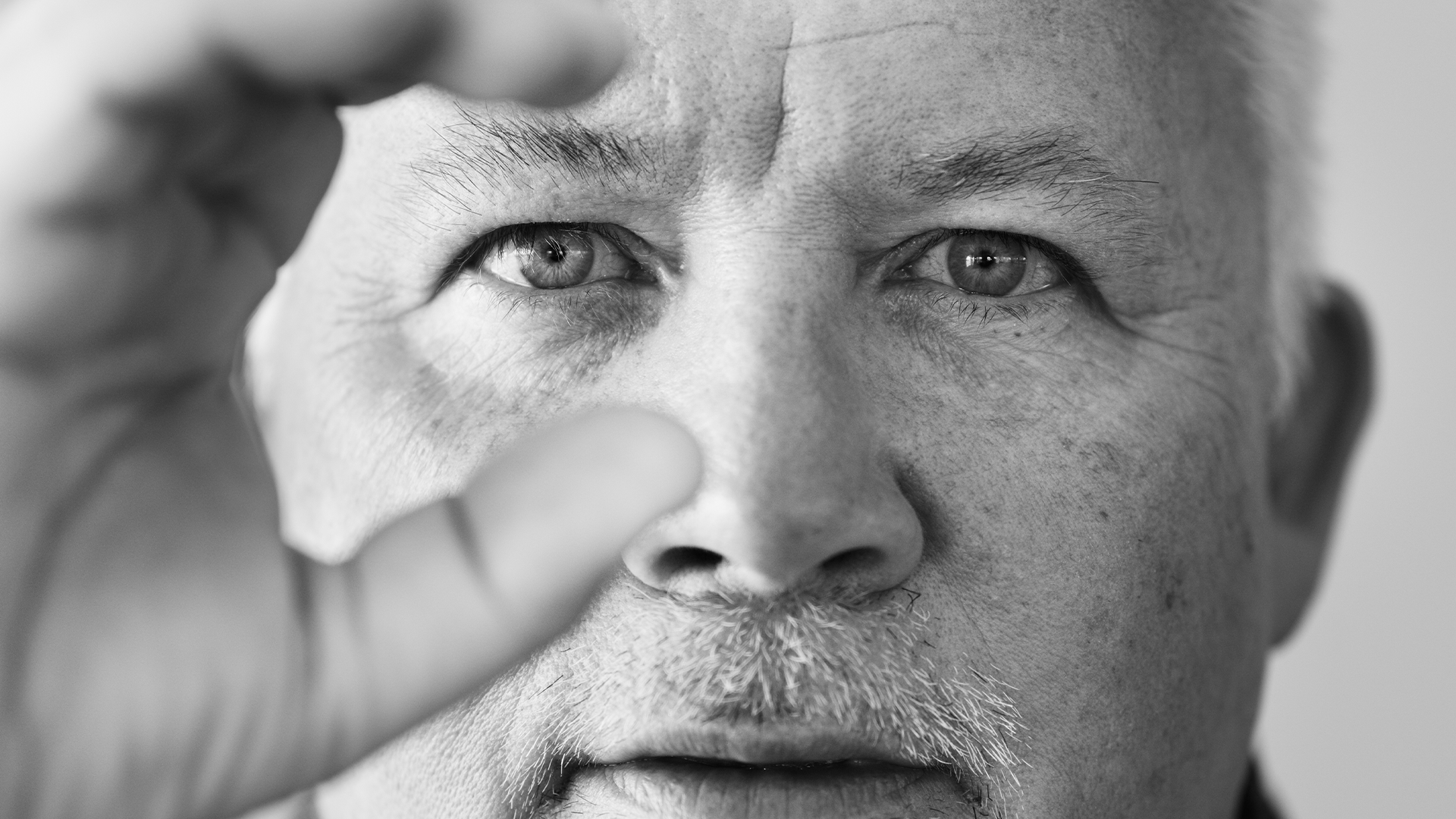 Kent Härstedt i en svartvit närbild. Han tittar in i kameran och håller upp händerna där fingrarna går i en halvcirkel runt hans högra öga.