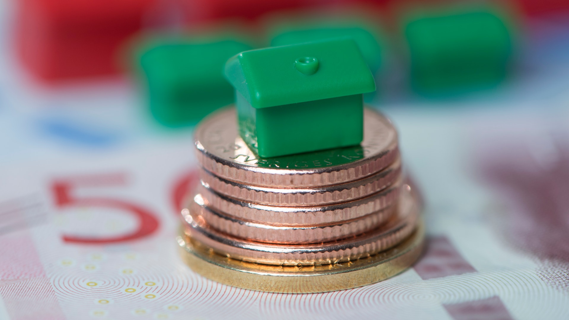 Litet grönt hus på en stapel mynt med sedlar i bakgrunden