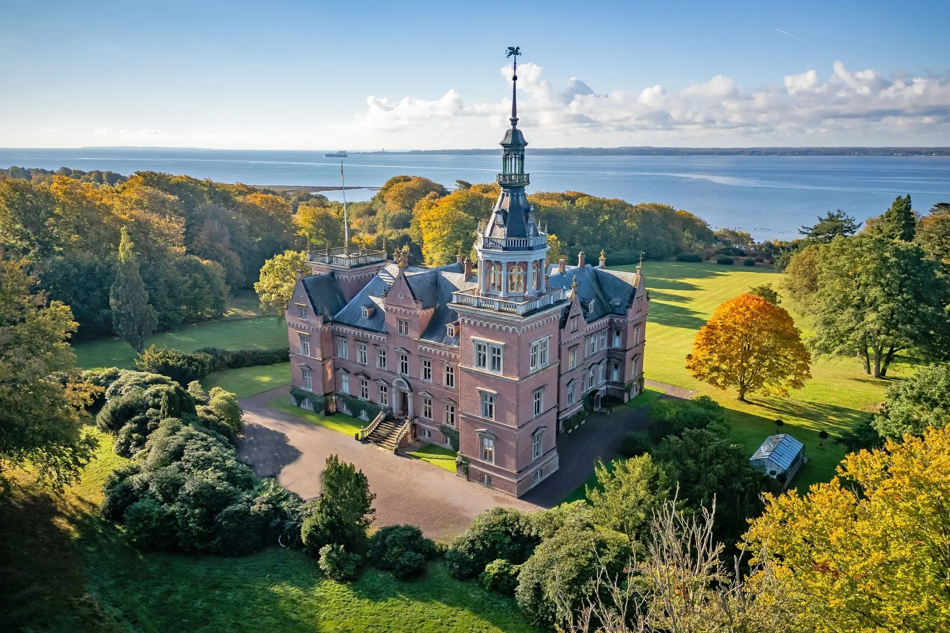 Rött tegelslott med torn omgivet av grön natur och skog, med vatten i bakgrunden