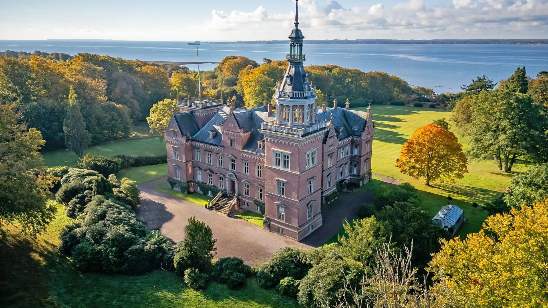 Rött tegelslott med torn omgivet av grön natur och skog, med vatten i bakgrunden