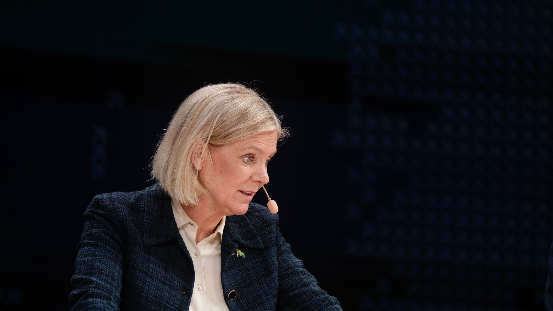 Kvinna i mörkblå kavaj sitter vid ett bord under en presentation och talar.