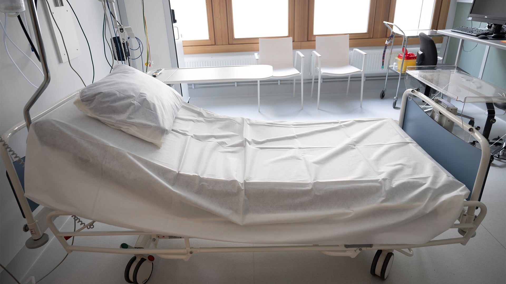 Sjukhussäng med vit bäddning i ett ljust patientrum med medicinsk utrustning och stora fönster.