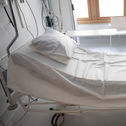 Sjukhussäng med vit bäddning i ett ljust patientrum med medicinsk utrustning och stora fönster.